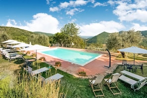 Zona de piscina con sillas de descanso y sombrillas, rodeada por un jardín ajardinado y colinas al fondo.