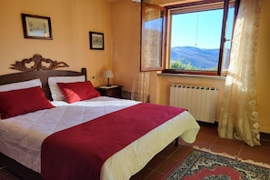 Una cama con ropa de cama roja y blanca, una mesita de noche con una lámpara, una ventana con vistas a colinas y un radiador.