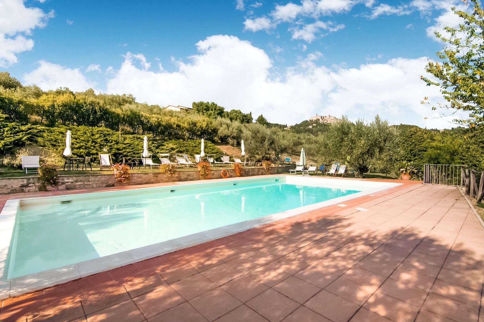 A rectangular swimming pool with a tiled deck, surrounded by lounge chairs and umbrellas, set against a backdrop of lush greenery and hills.