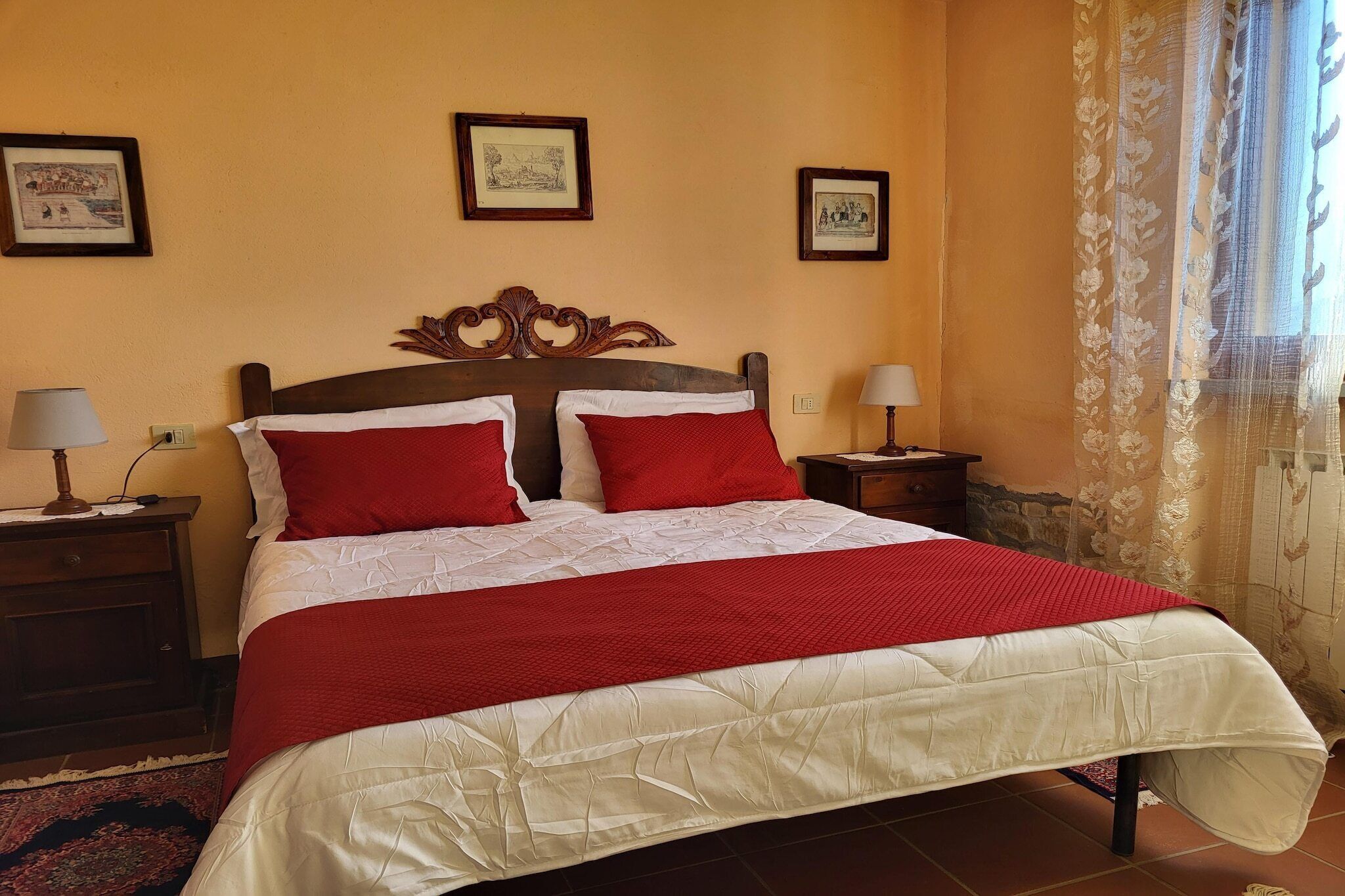 Una cama con cabecera de madera, dos almohadas rojas y colcha blanca. Hay dos mesitas de noche con lámparas, un cuadro enmarcado en la pared y una ventana con cortina estampada.