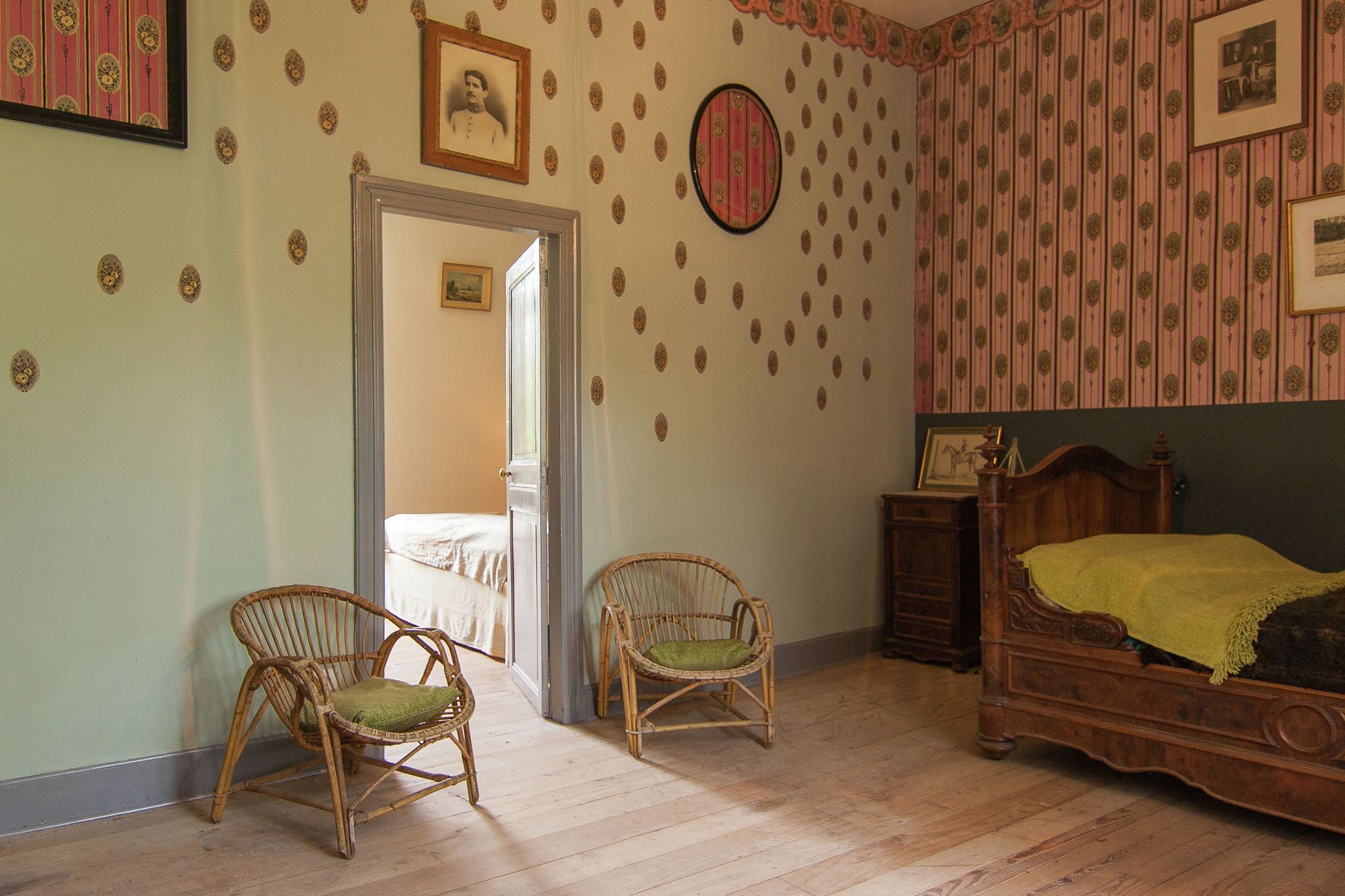 Una habitación con una cama de madera, dos sillas de mimbre, un aparador y una puerta que da a otra habitación.