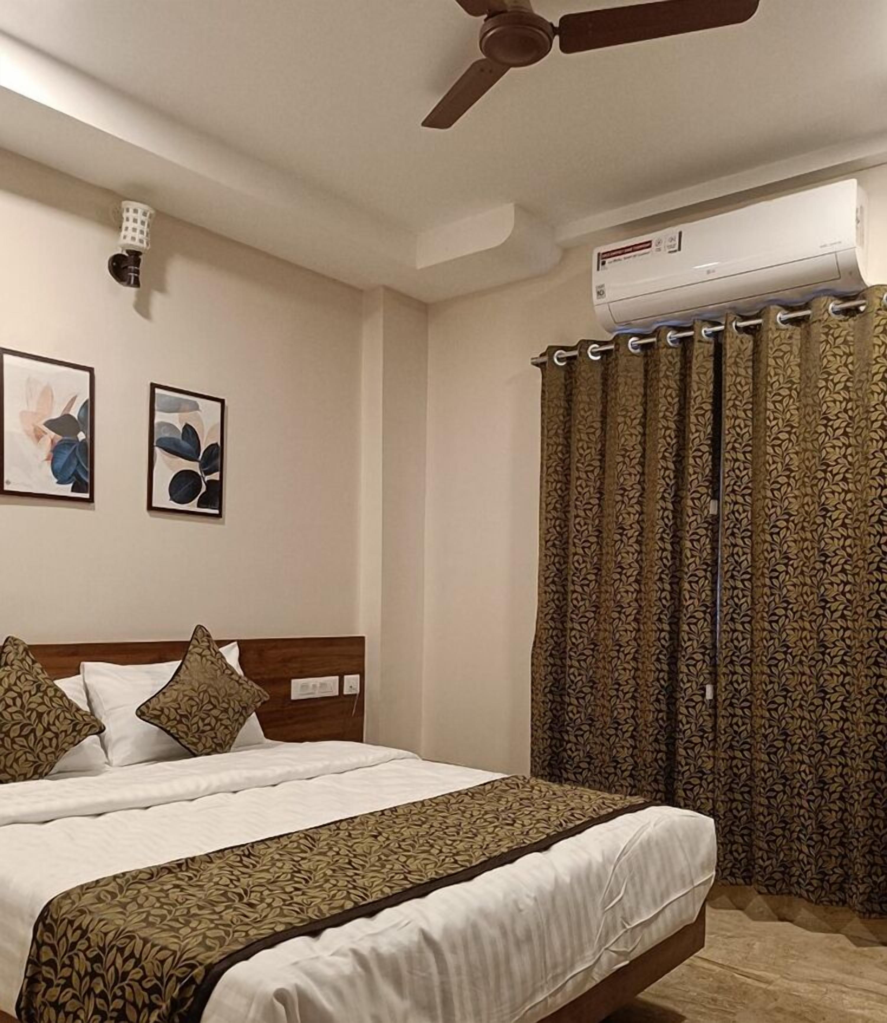 A bedroom with a bed, a ceiling fan, an air conditioning unit, patterned curtains, and two framed artworks on the wall.
