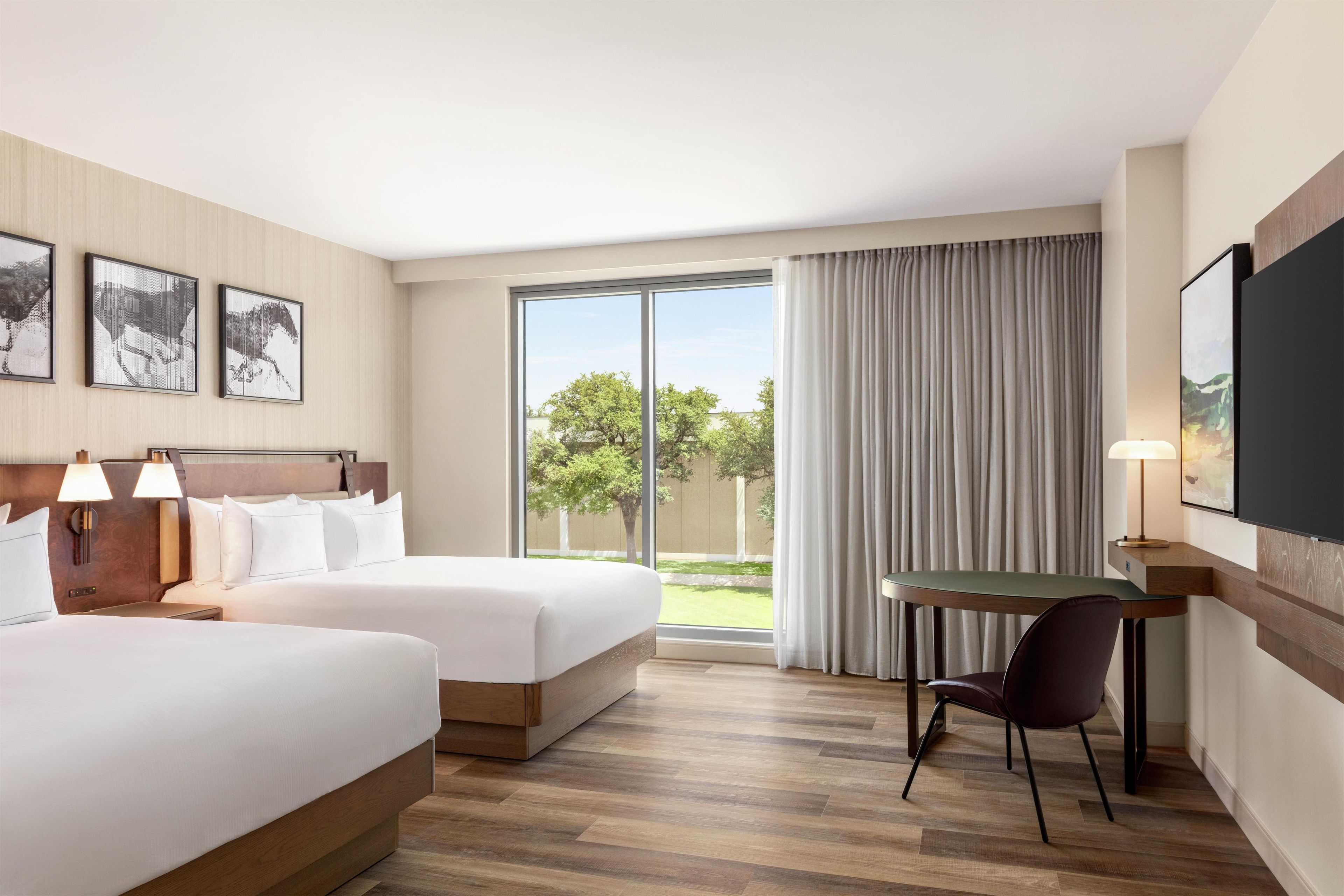 A hotel room with two beds, a desk with a chair, a large window with curtains, and artwork on the walls.