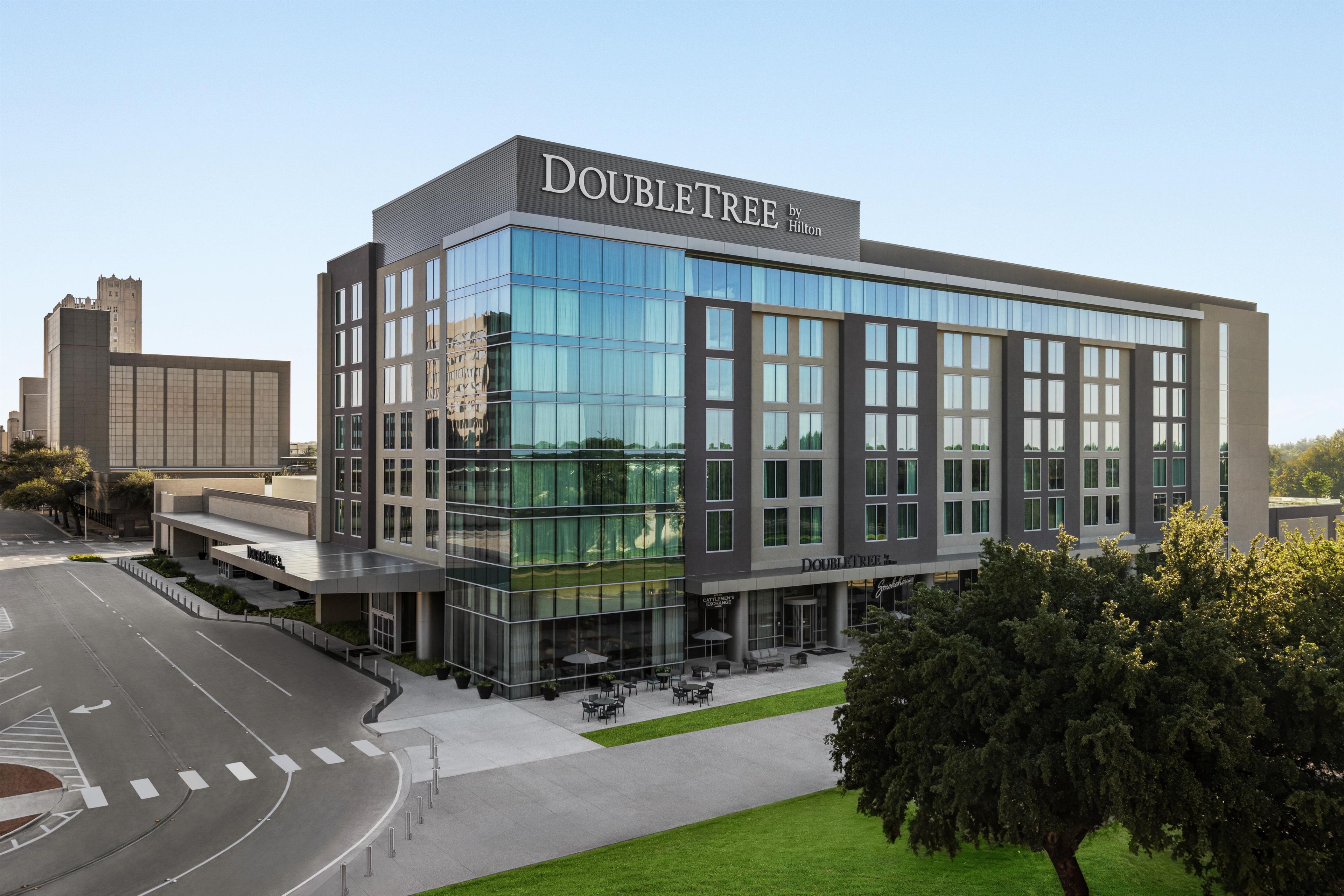 Un hôtel moderne à la façade en verre, appelé « DOUBLETREE by Hilton », qui dispose d’un vaste espace extérieur aménagé et d’une entrée imposante.