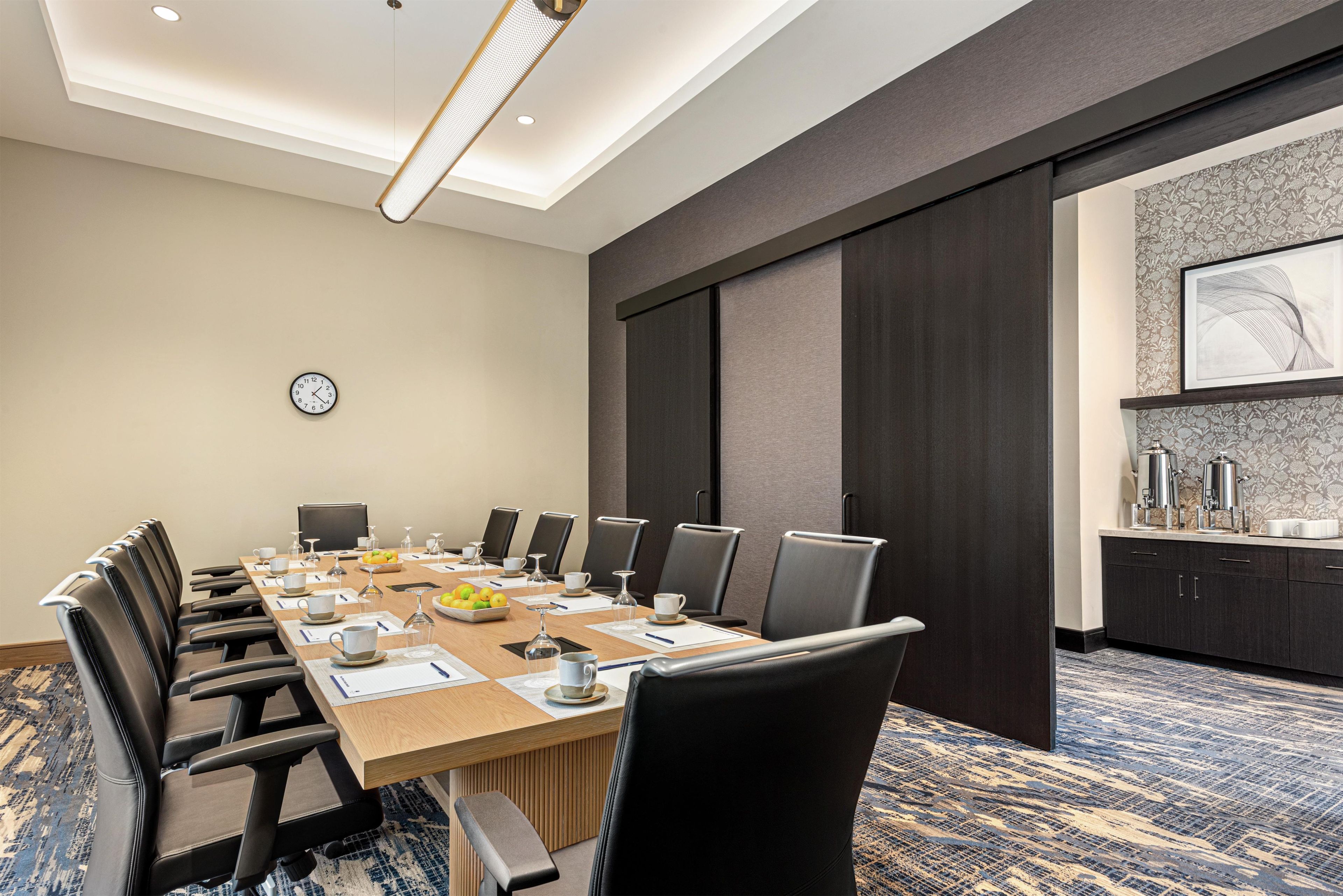A conference room with a long table, chairs, a clock, and a coffee station.