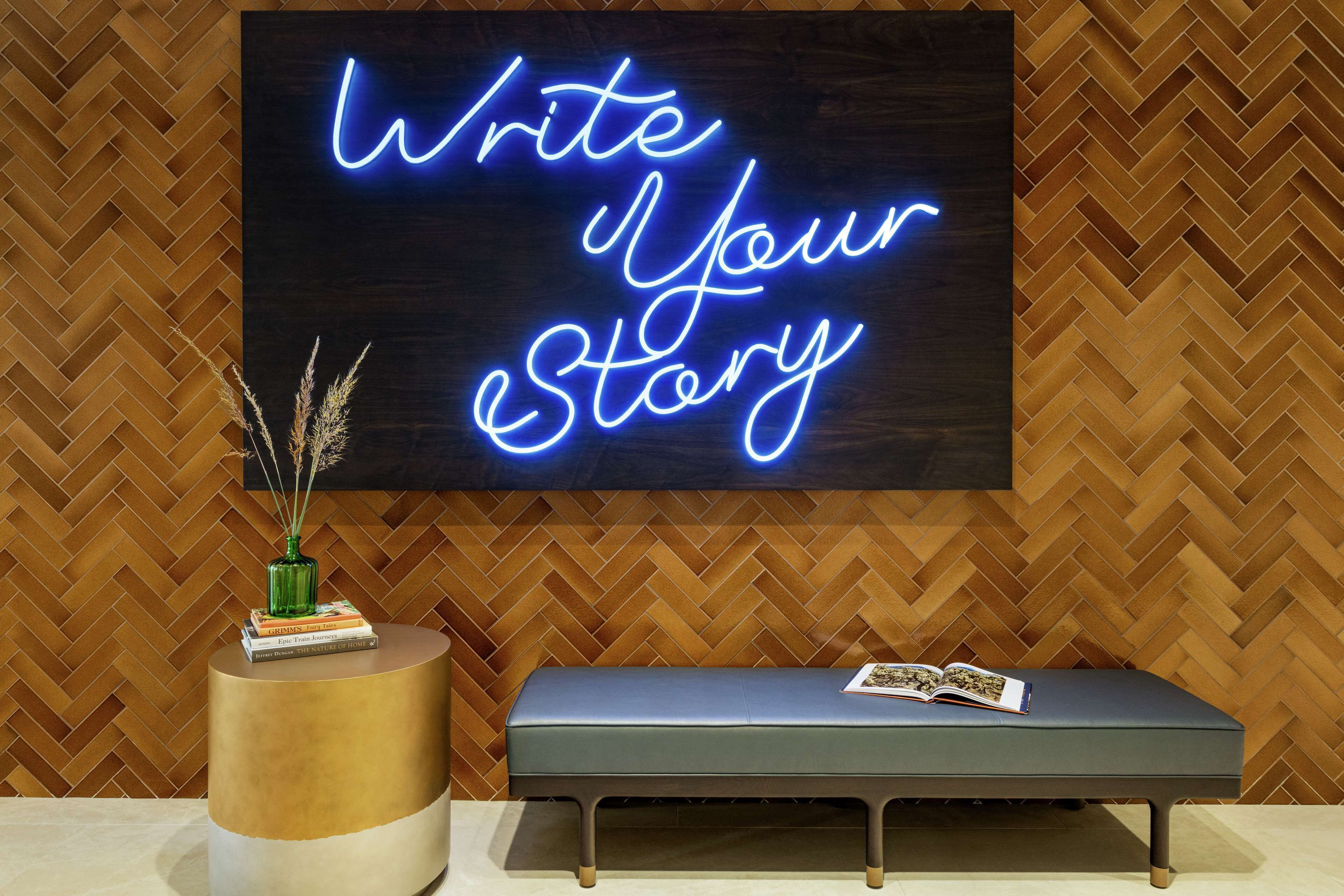 A room with a herringbone-patterned wooden wall, a neon sign that reads "Write Your eStory," a round gold table with a green vase and books, and a bench with an open book.