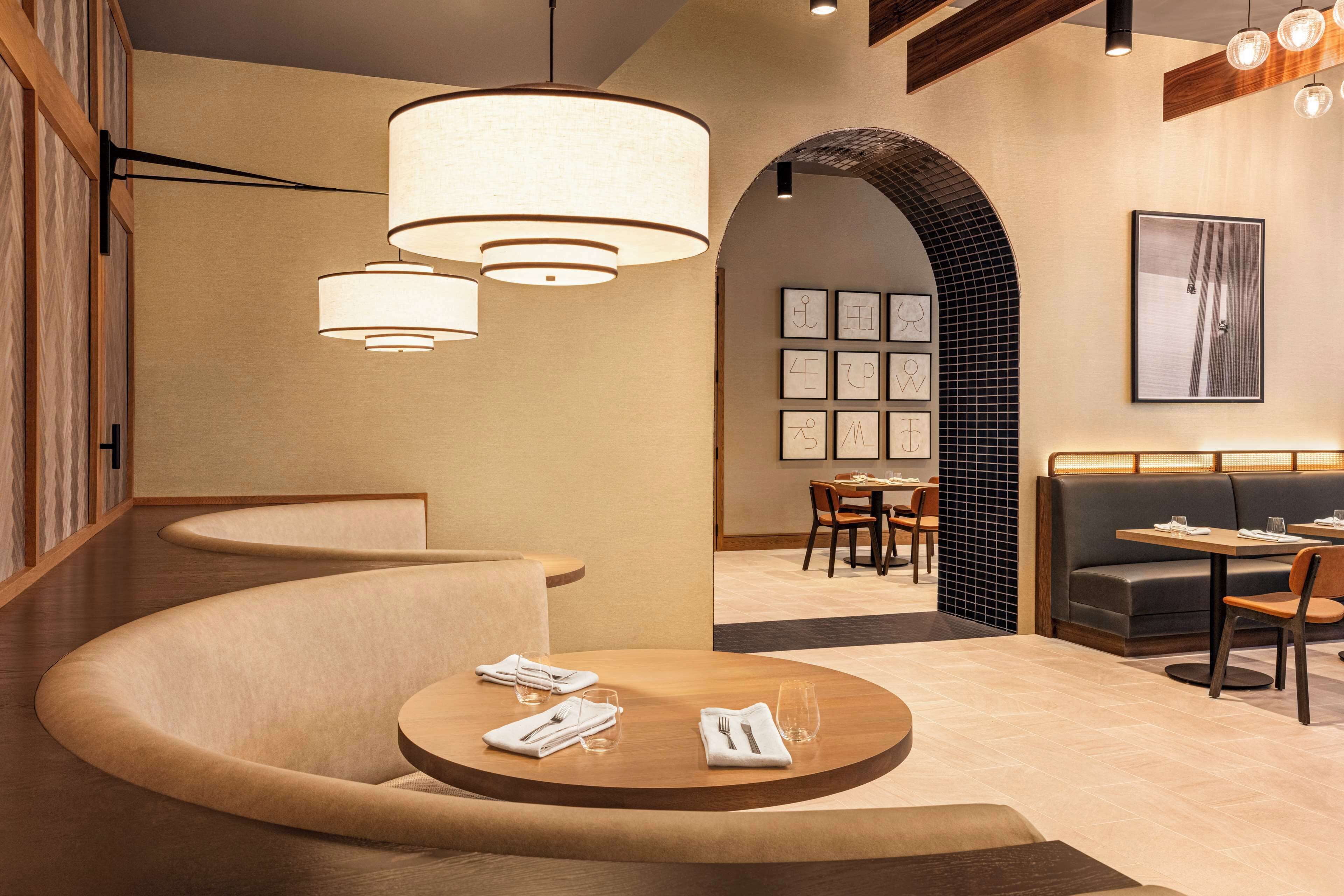 Et moderne restaurantinteriør med et buet siddeområde, flere spiseborde og en blanding af træ- og polstrede møbler.