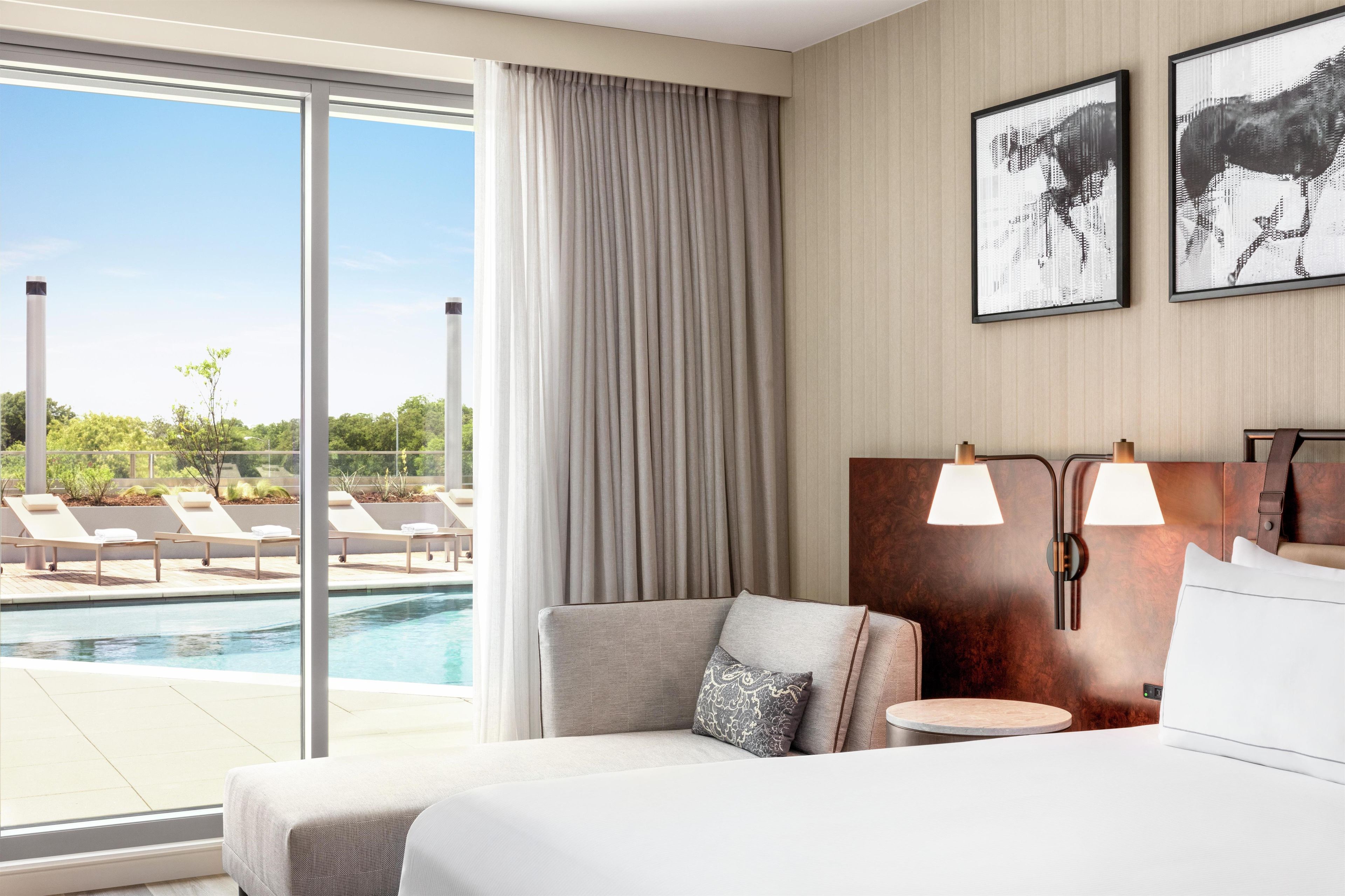 Habitación de hotel con un ventanal grande, una cama, una mesita de noche, una lámpara y vistas a una piscina y sillas de descanso.