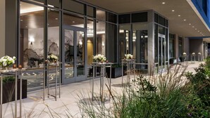 Un hôtel moderne avec une façade élégante, de grandes portes vitrées, des tables en plein air agrémentées de compositions florales et un hall d’entrée bien éclairé.