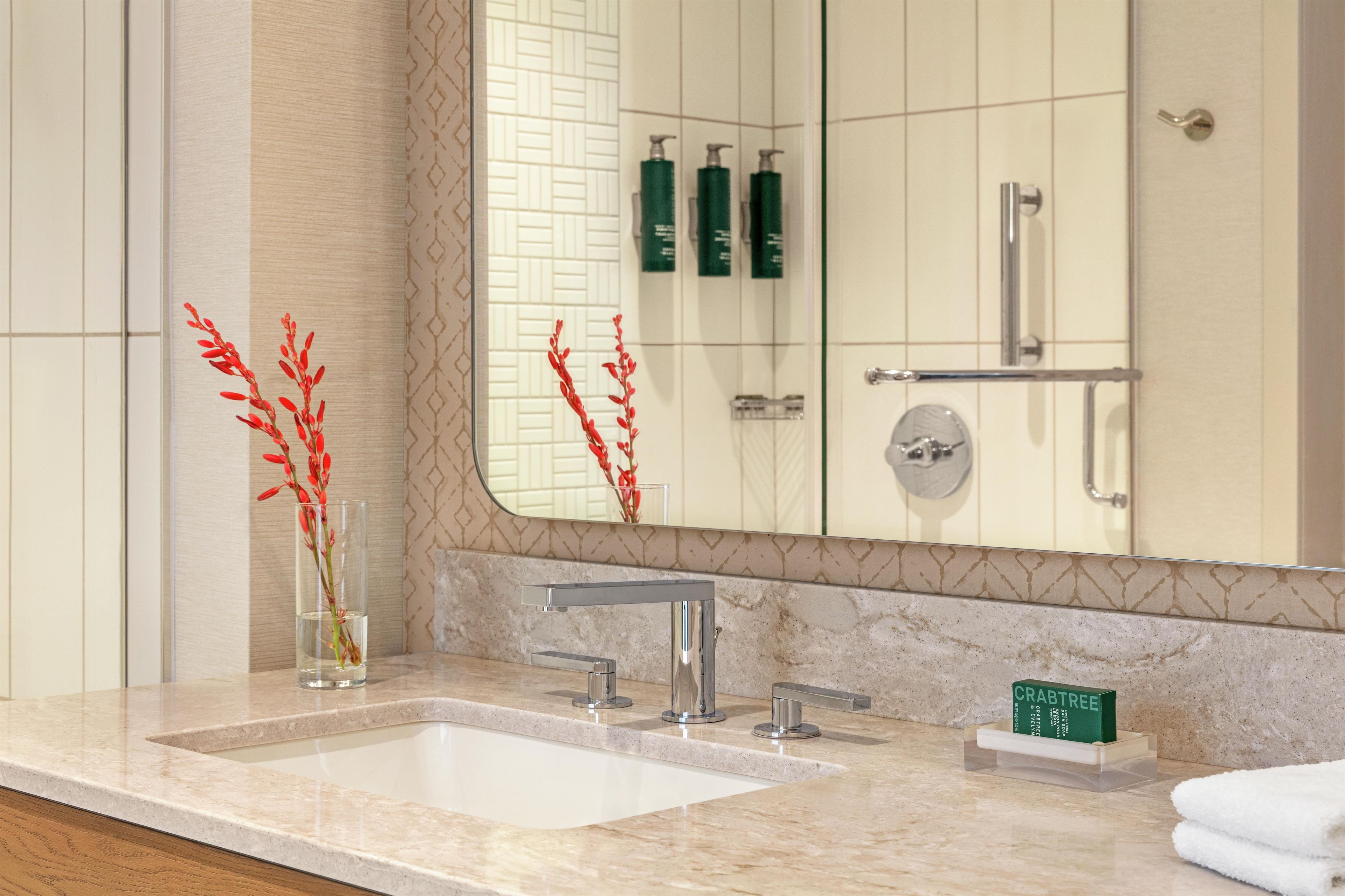 A bathroom with a marble sink, a mirror, and a glass vase with red flowers.