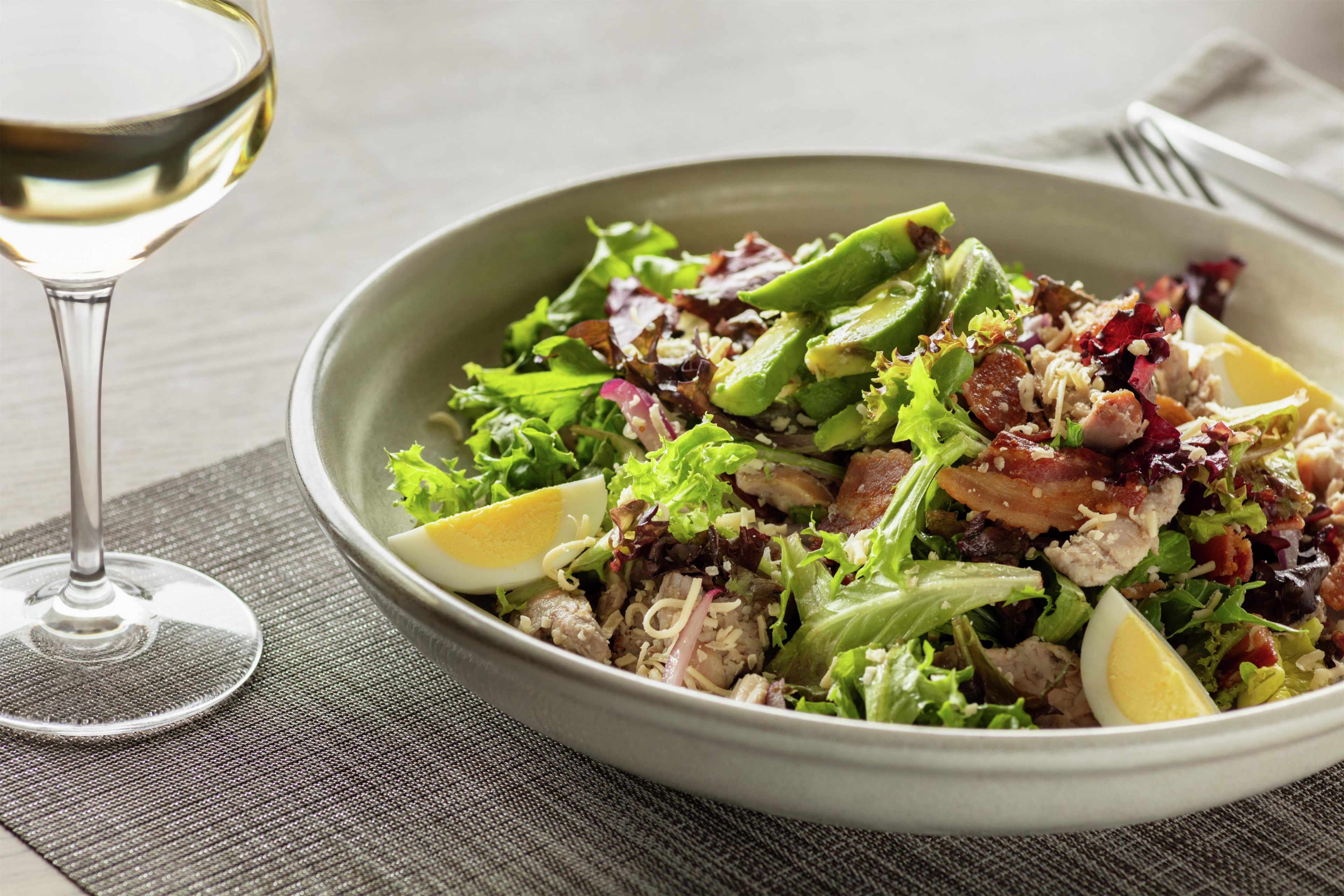 A bowl of salad with various ingredients and a glass of white wine on a table.