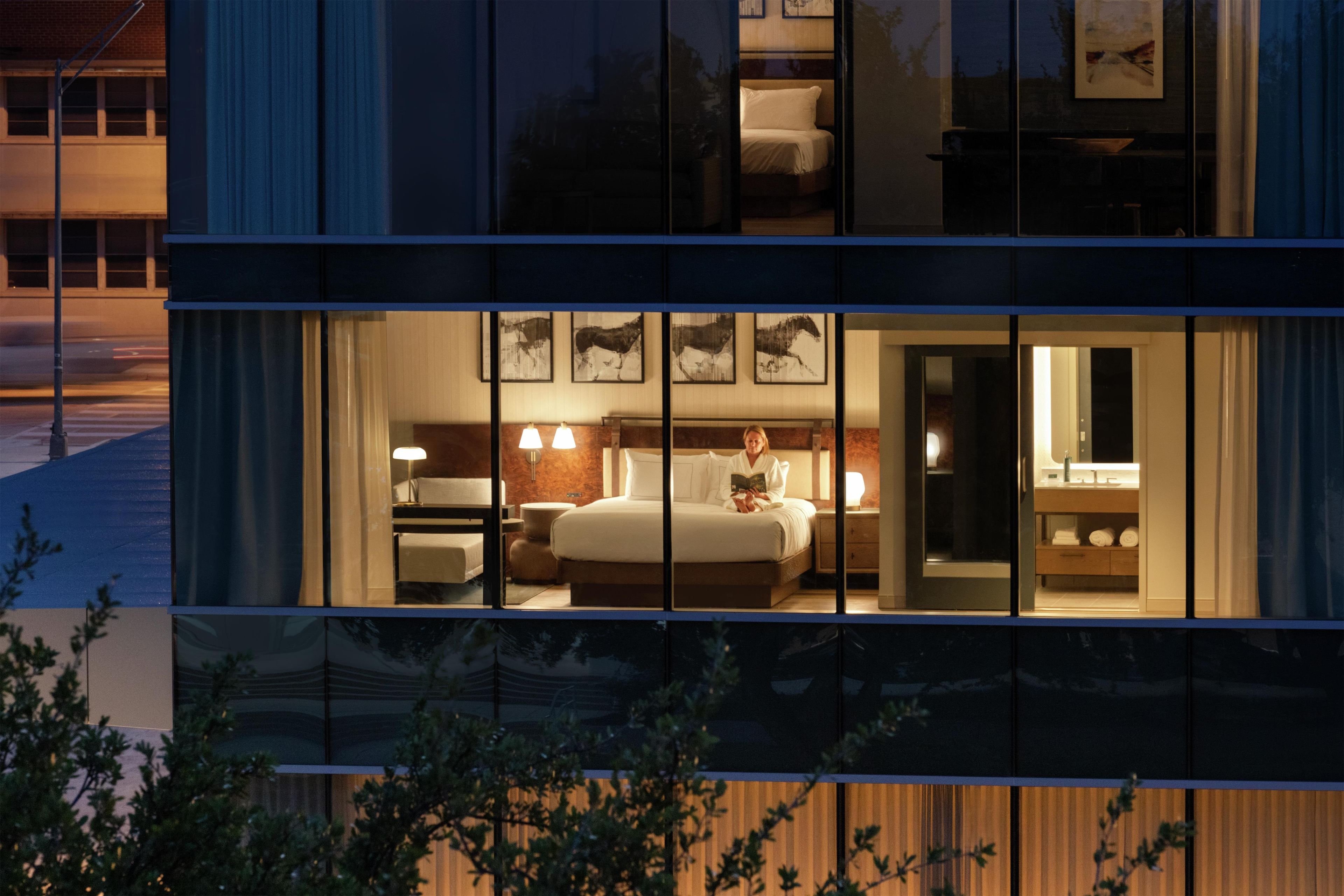 A modern hotel room with a large bed, bedside tables, and a bathroom visible through the glass wall.