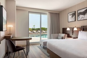 A hotel room with a large bed, a desk, a chair, a small table, and a view of a swimming pool.