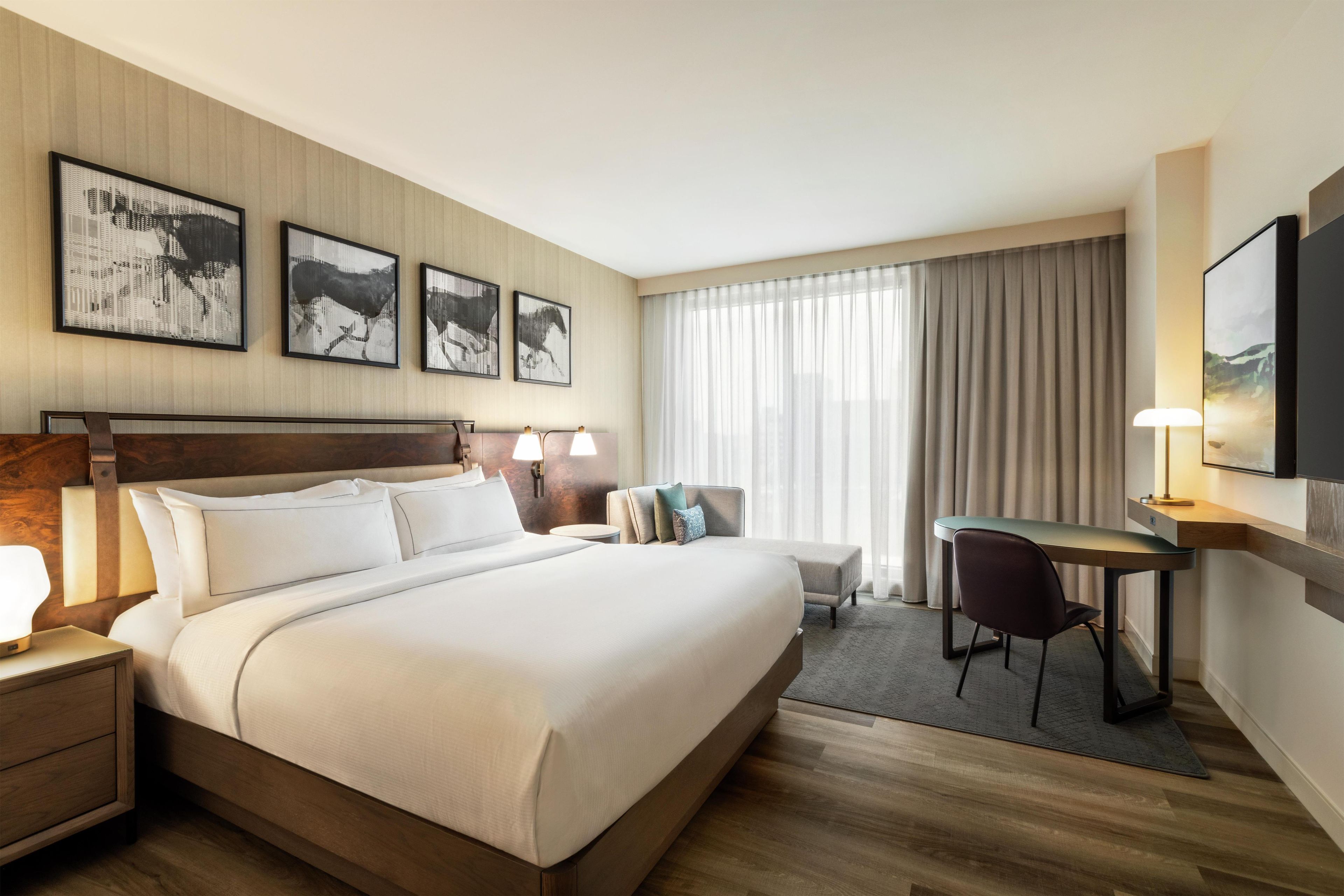 Habitación de hotel con una cama grande, un escritorio, una silla y un televisor. Hay cuadros en la pared y una ventana con cortinas.