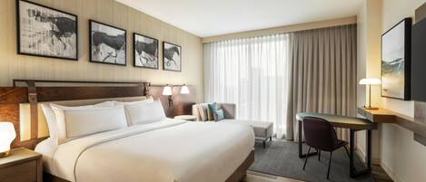 Habitación de hotel con una cama grande, un escritorio, una silla y un televisor. Hay cuadros en la pared y una ventana con cortinas.