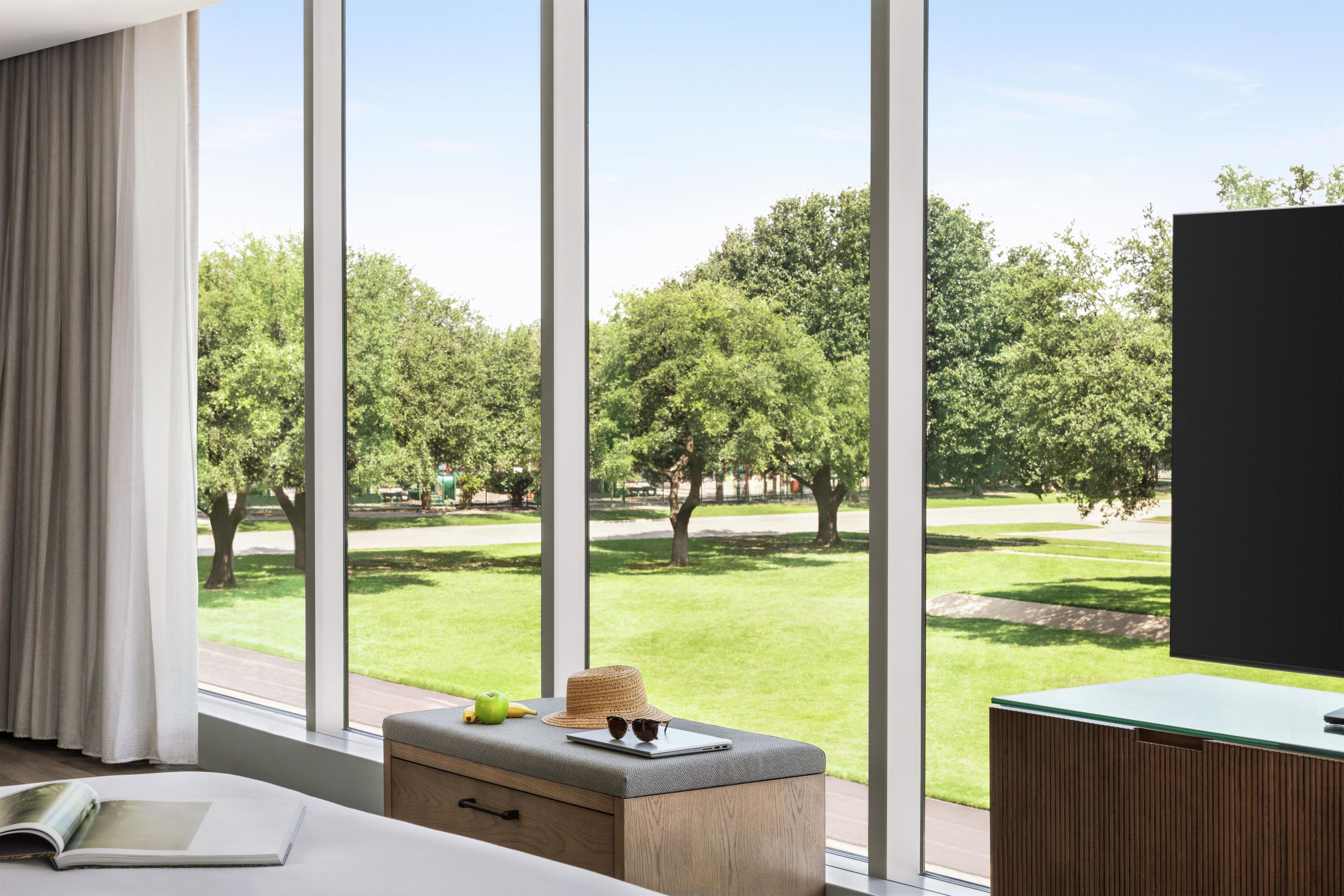 A modern hotel room with a large window, a wooden bench, a flat-screen TV, and a view of a park with trees and grass.
