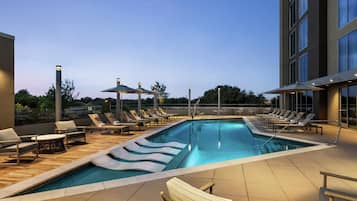 Zona de piscina en la azotea con sillones de descanso y sombrillas, rodeada por un edificio moderno.