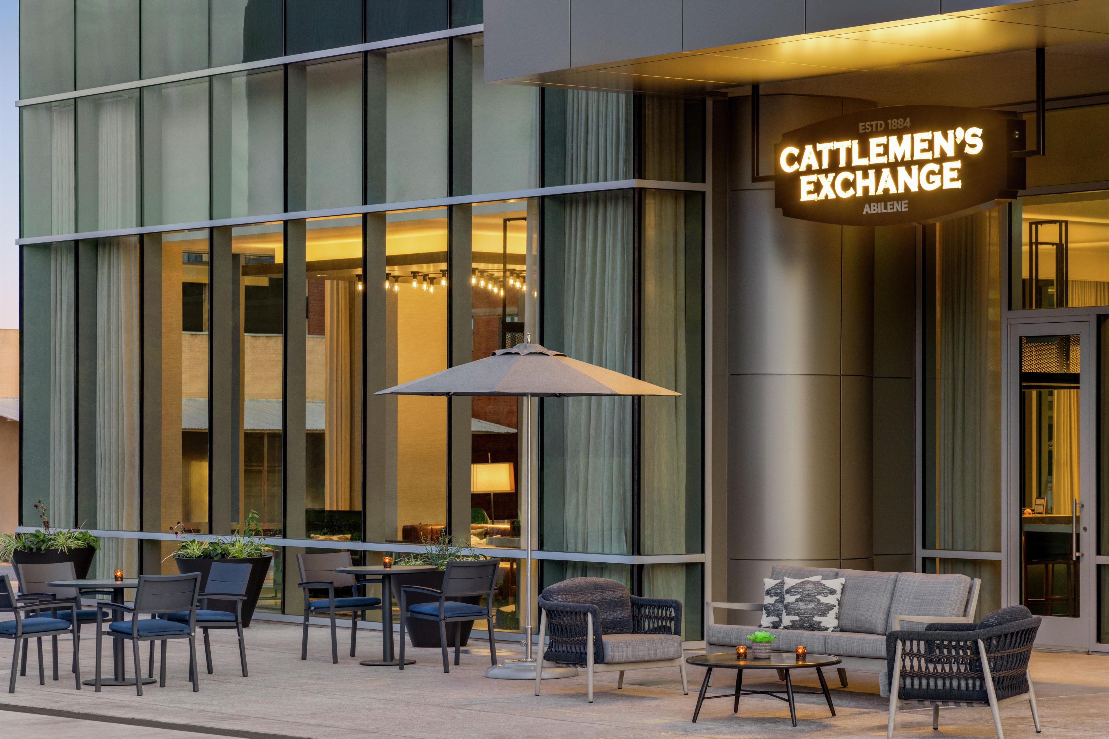An outdoor seating area with tables and chairs, a large umbrella, and a building with a sign reading 'Cattlemen's Exchange.'