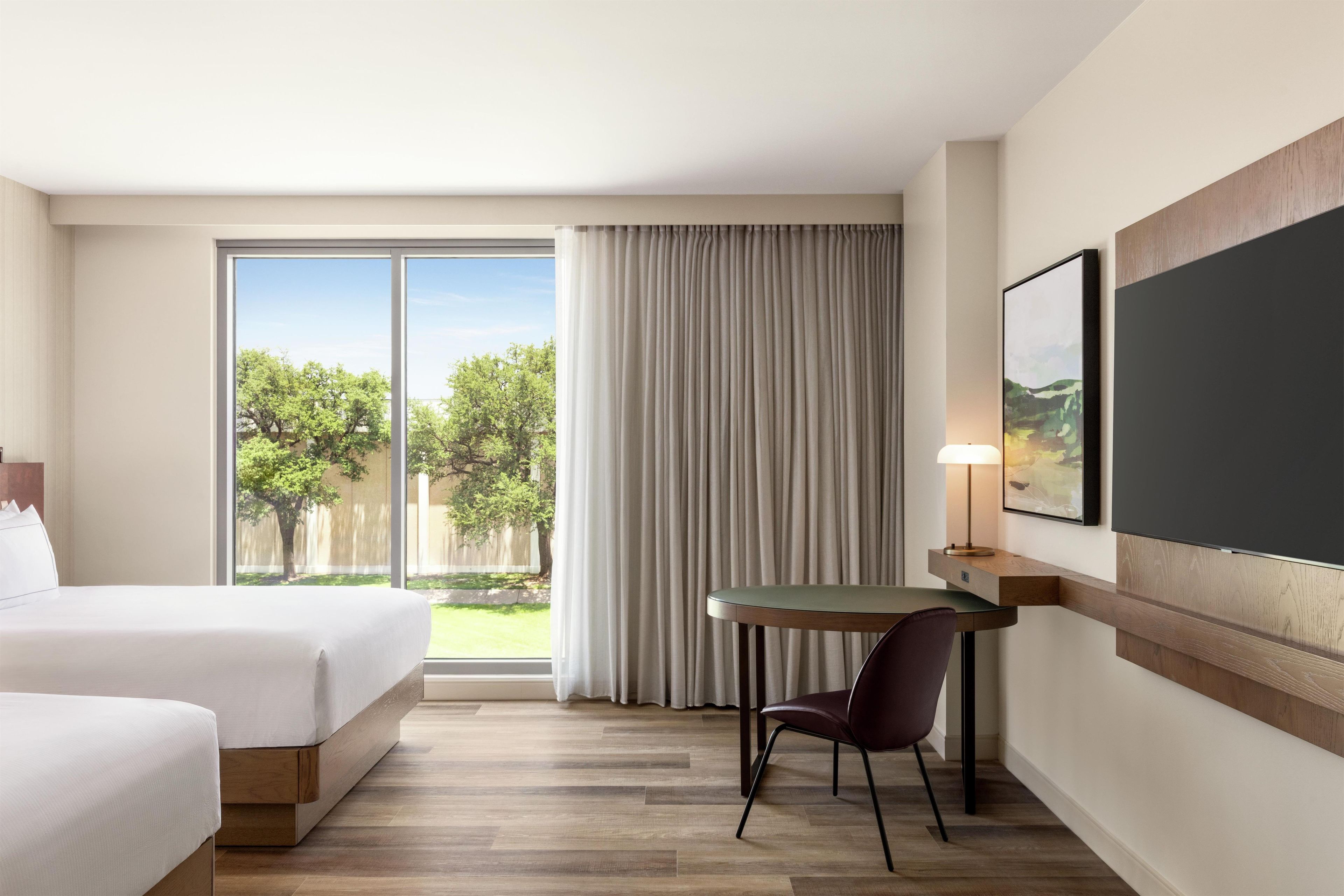 Habitación de hotel con cama, escritorio, silla, lámpara y ventana grande que ofrece vistas a árboles y una valla.