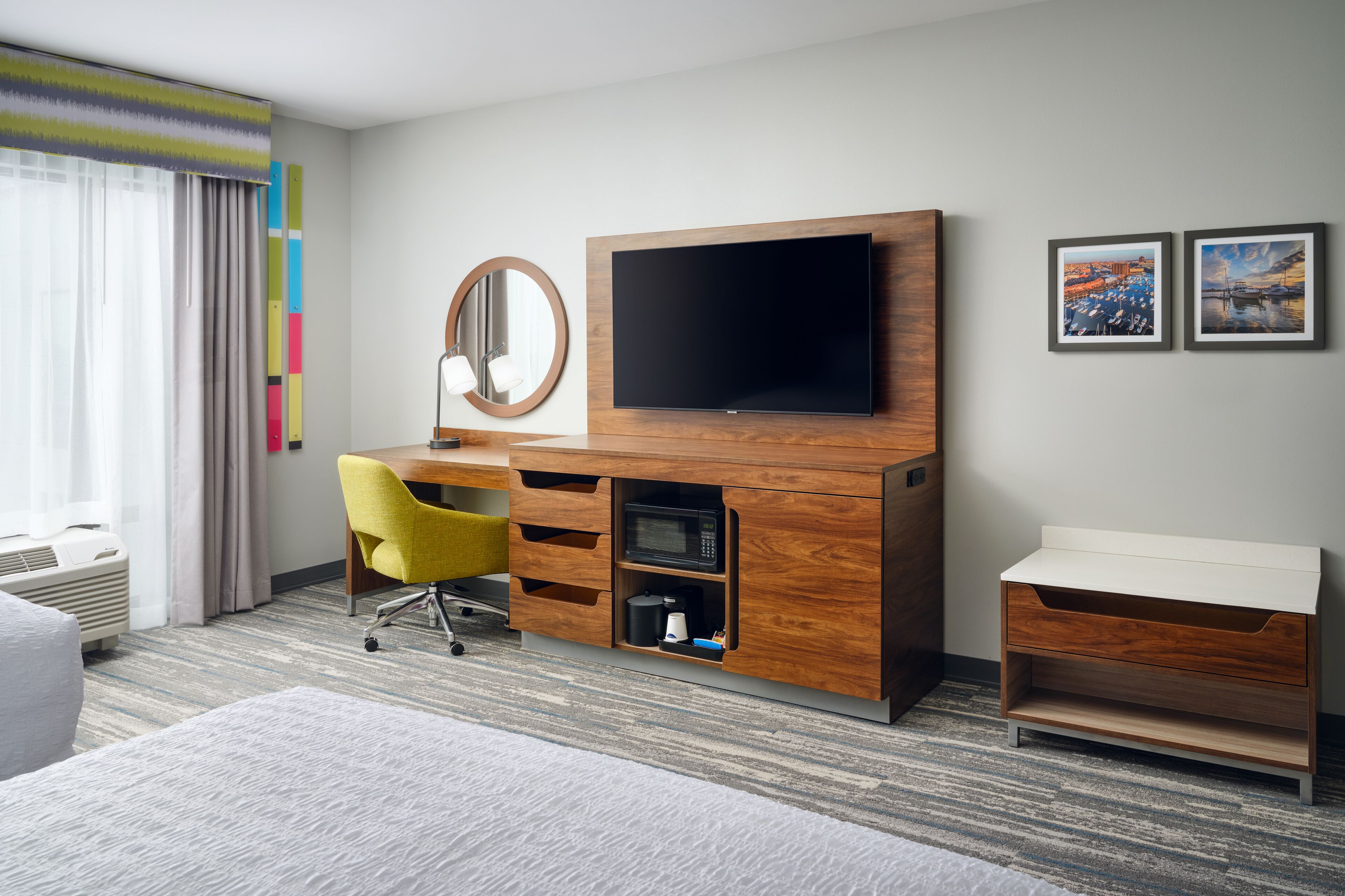 A hotel room with a wooden desk, a flat-screen TV, a mirror, and two framed pictures on the wall.