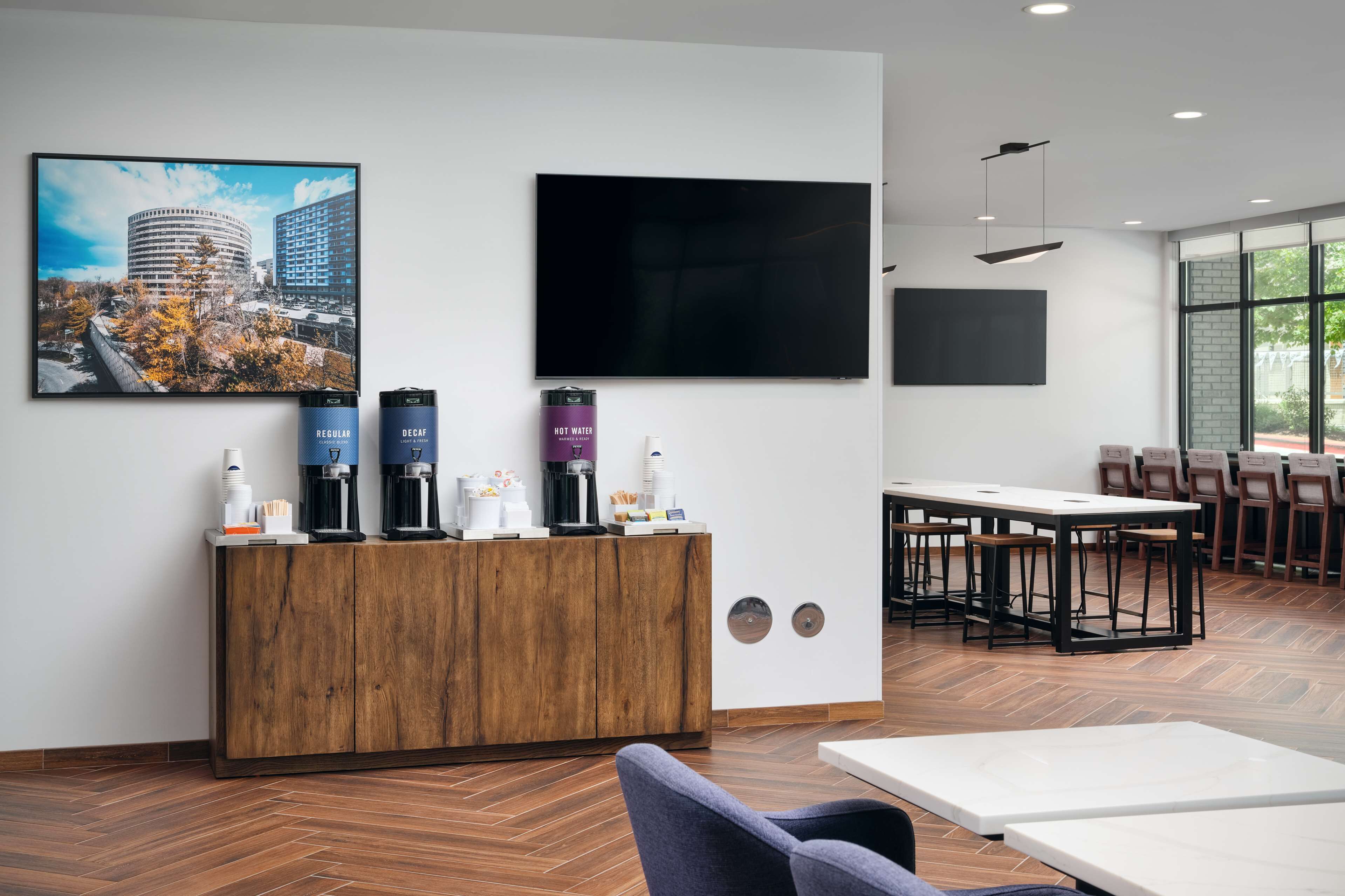 A modern hotel lobby with a wooden reception desk, a large flat-screen TV, and a seating area with tables and chairs.