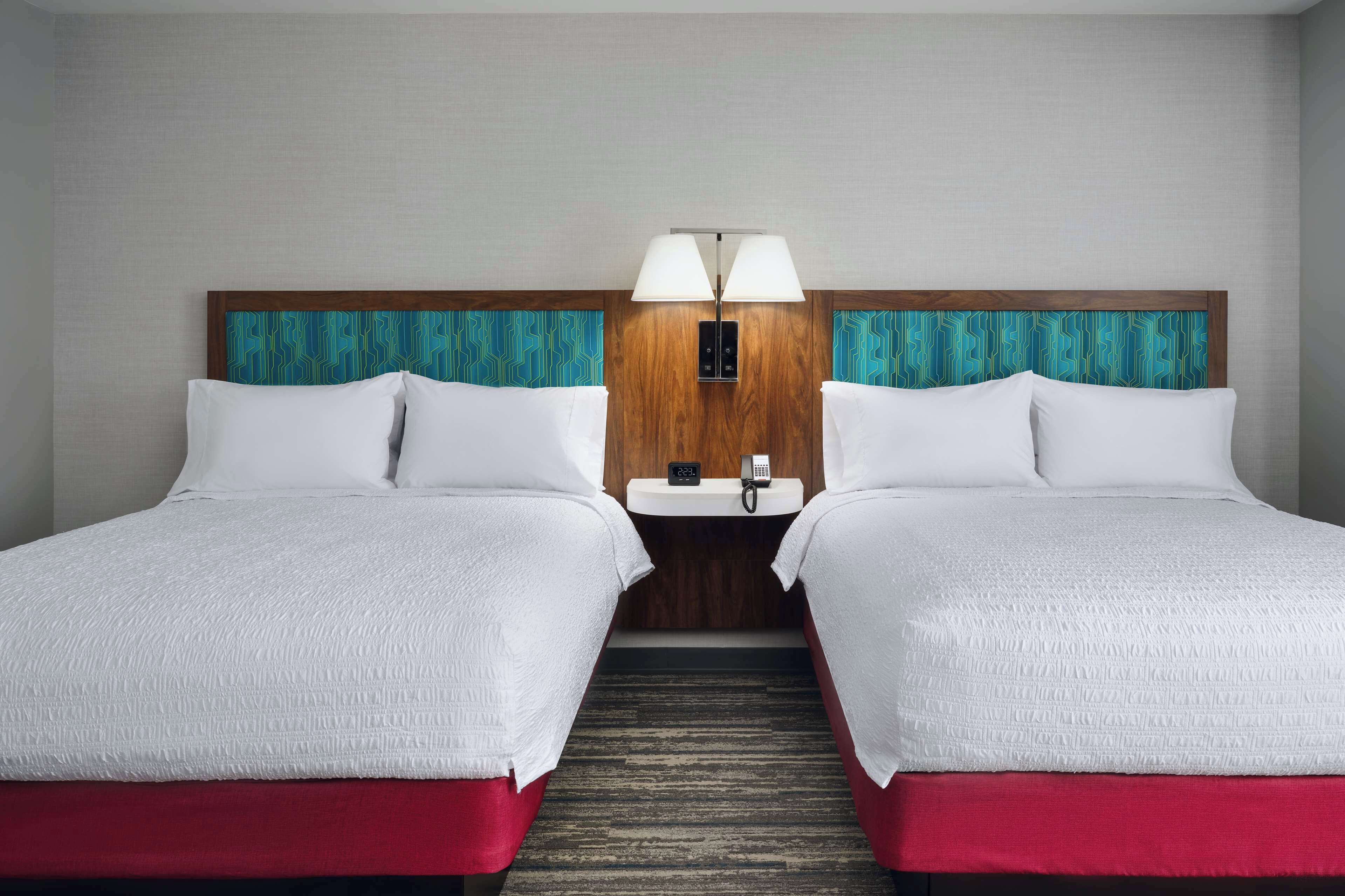 A hotel room with two beds, a wooden headboard, a nightstand with a phone and lamp, and a red carpet.