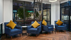 A seating area with blue patterned chairs and mustard-colored cushions, large windows with a view of the outside, and a modern ceiling light fixture. - Hampton Inn Towson Downtown (Towson)