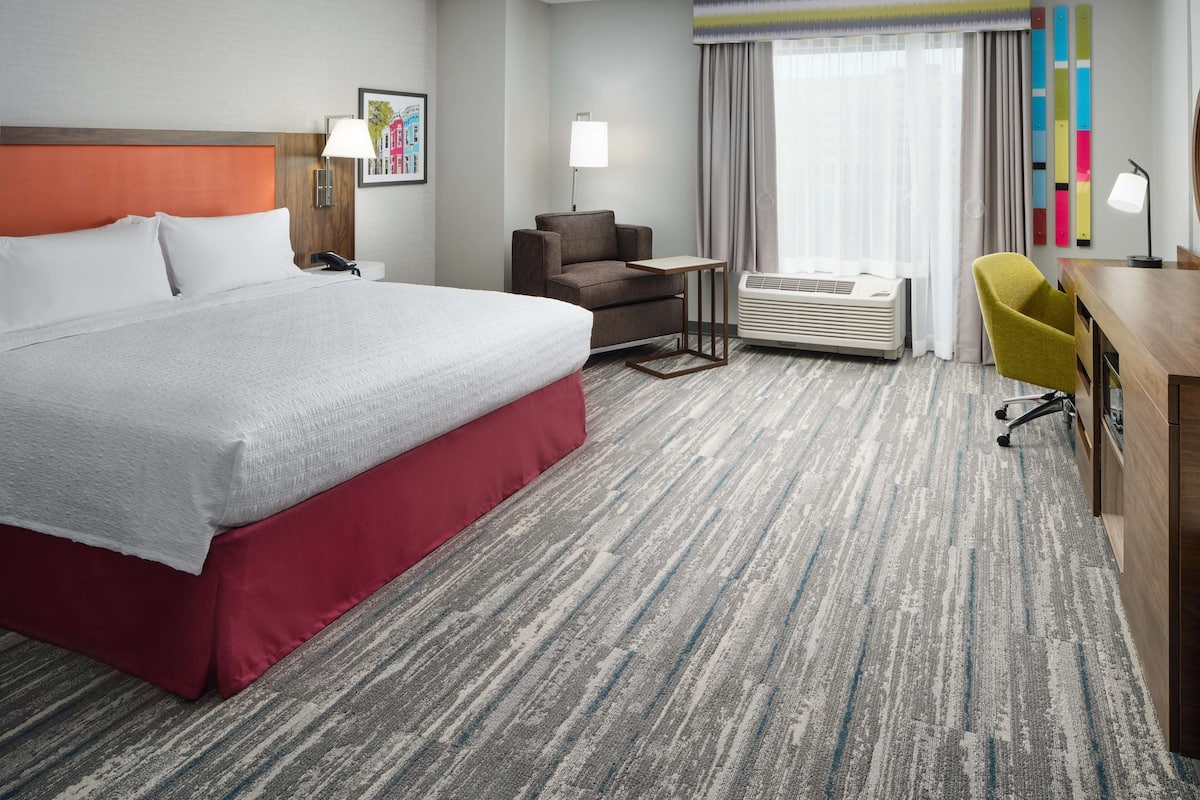 A hotel room with a large bed, a desk, a chair, a small table, an air conditioner, and a window with curtains.