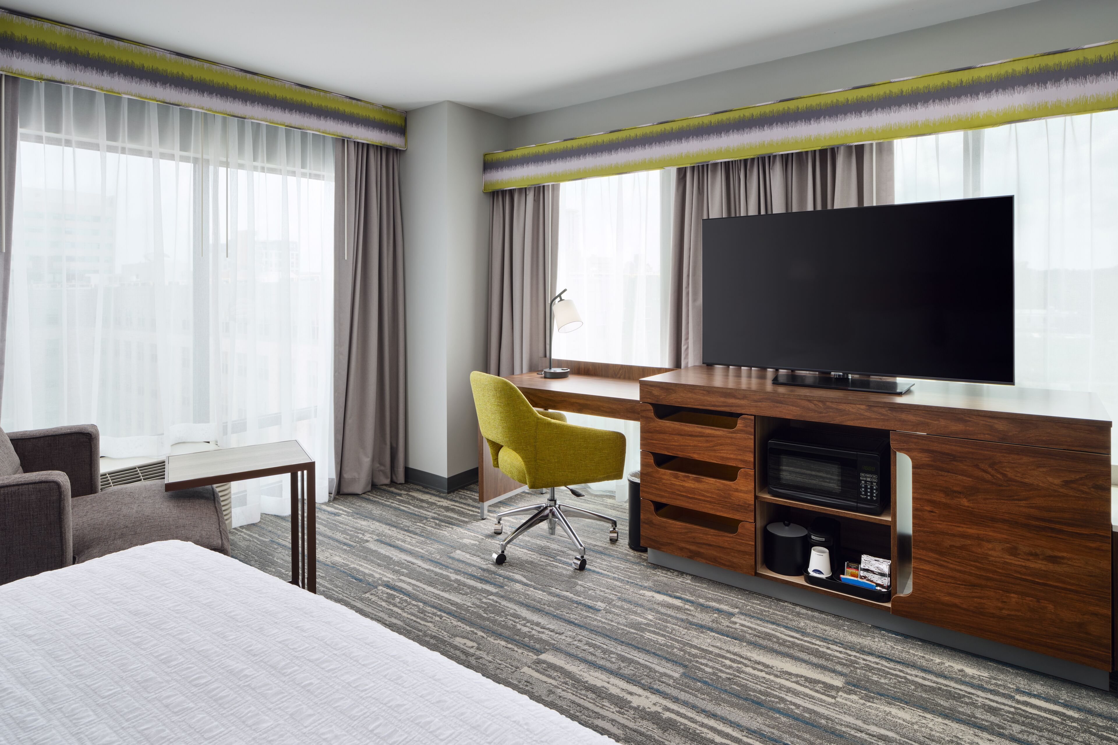 A hotel room with a large bed, a desk with a television, a yellow chair, and a minibar.