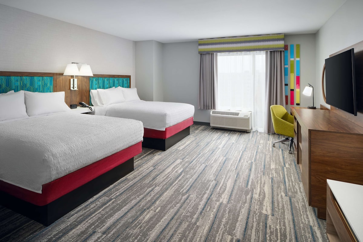 A hotel room with two beds, a desk, a chair, a TV, and a window with curtains.