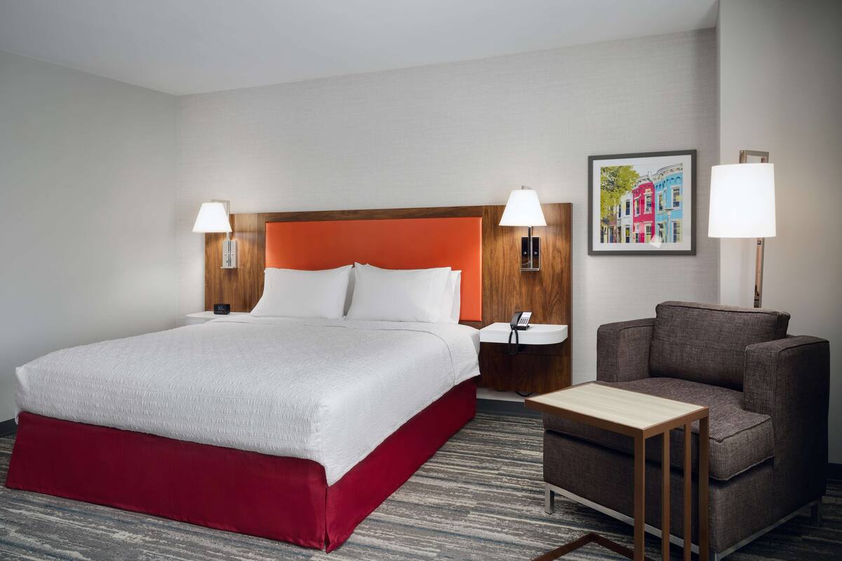 A hotel room with a large bed, a wooden headboard, two bedside lamps, a small table, a chair, and a framed picture on the wall.