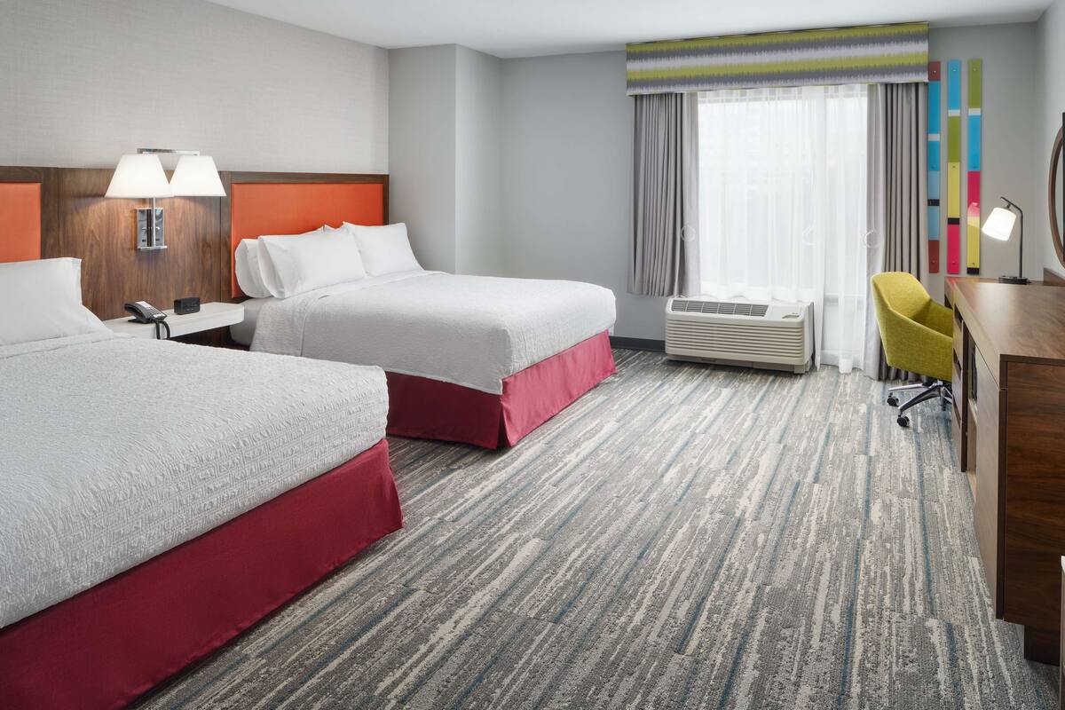 A hotel room with two beds, a desk, a chair, a phone, an air conditioner, and a window with curtains.
