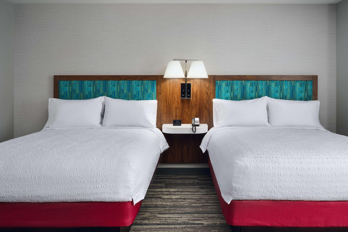 A hotel room with two beds, a wooden headboard, a bedside table with a lamp, and a clock.