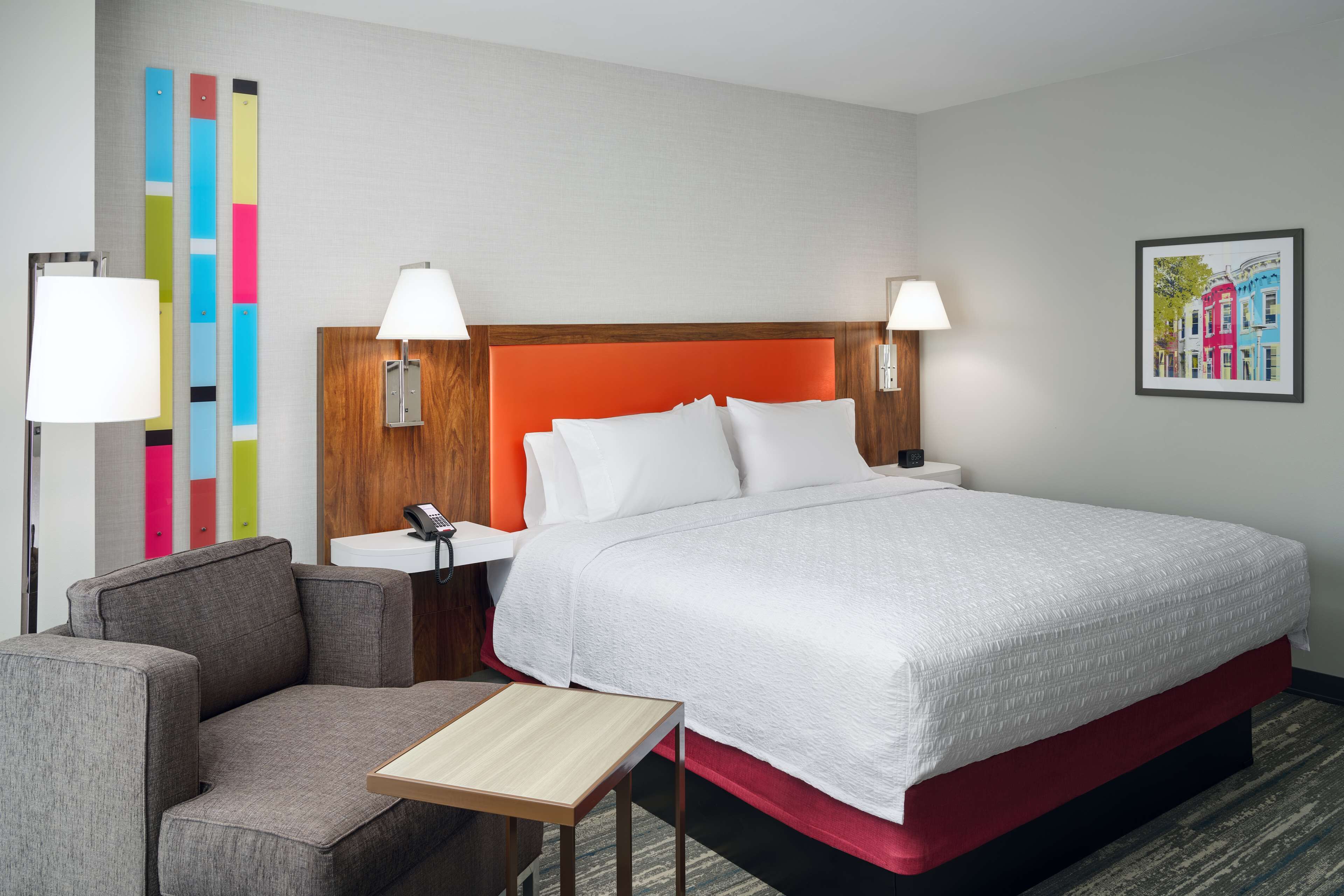 A hotel room with a large bed, two bedside lamps, a small table, a sofa, and a colorful wall decoration.