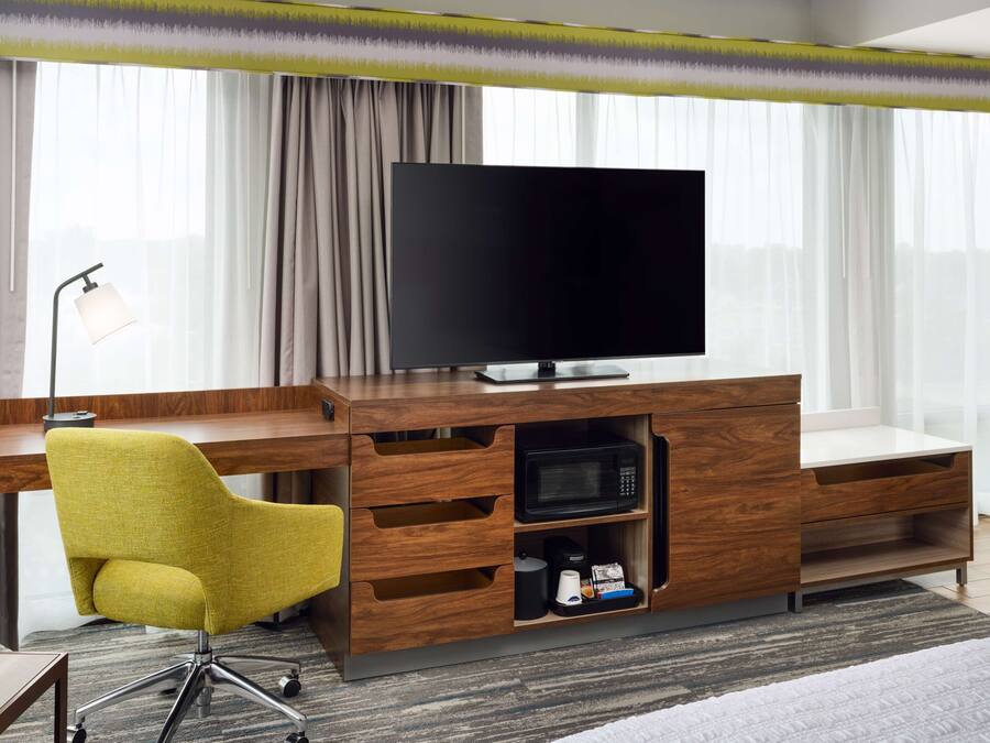 A modern hotel room with a wooden desk, a flat-screen TV, a yellow chair, a microwave, and a coffee maker.