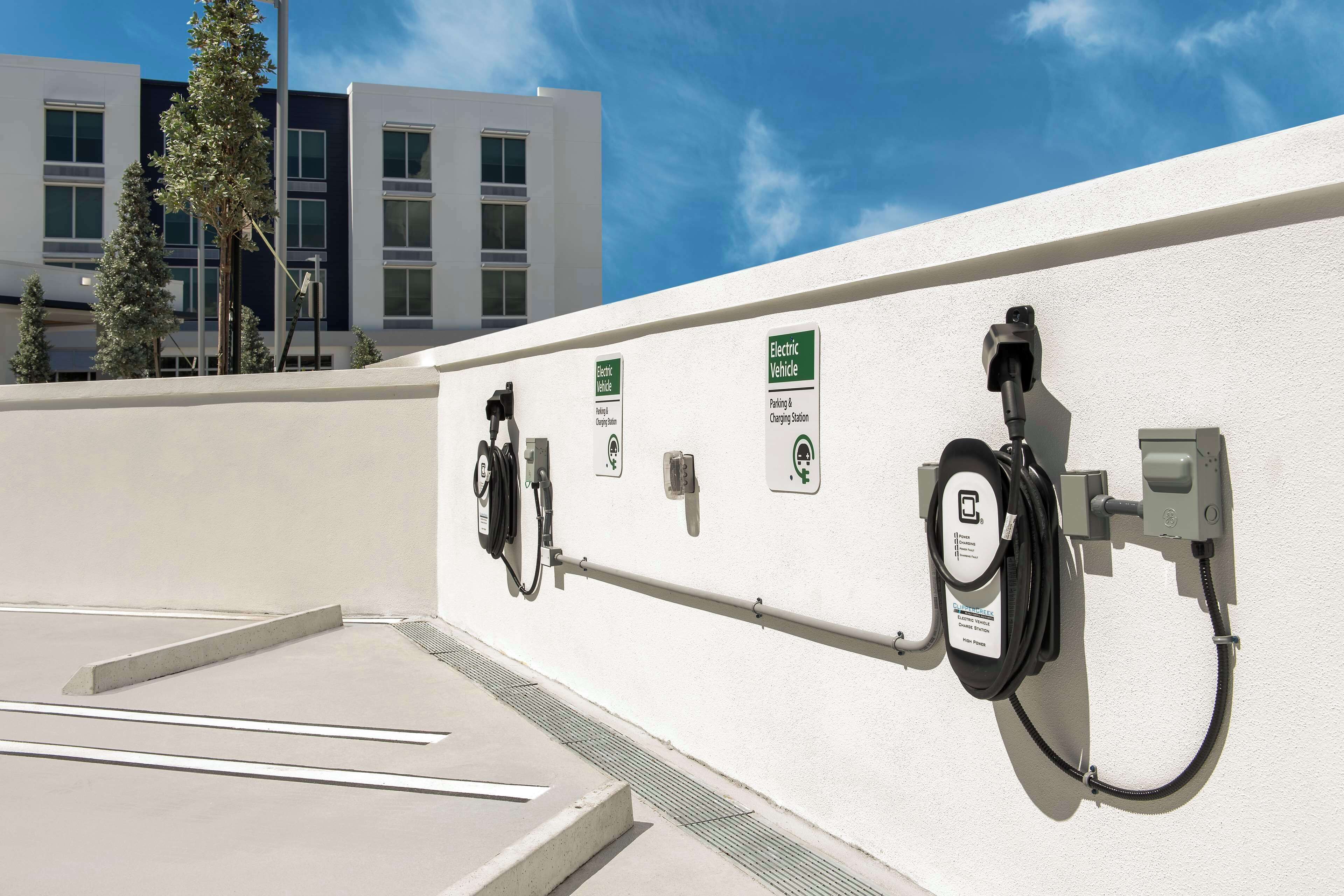 Uma estação de carregamento para veículos elétricos com cabo e tomada, um sinal de estacionamento e um edifício ao fundo.