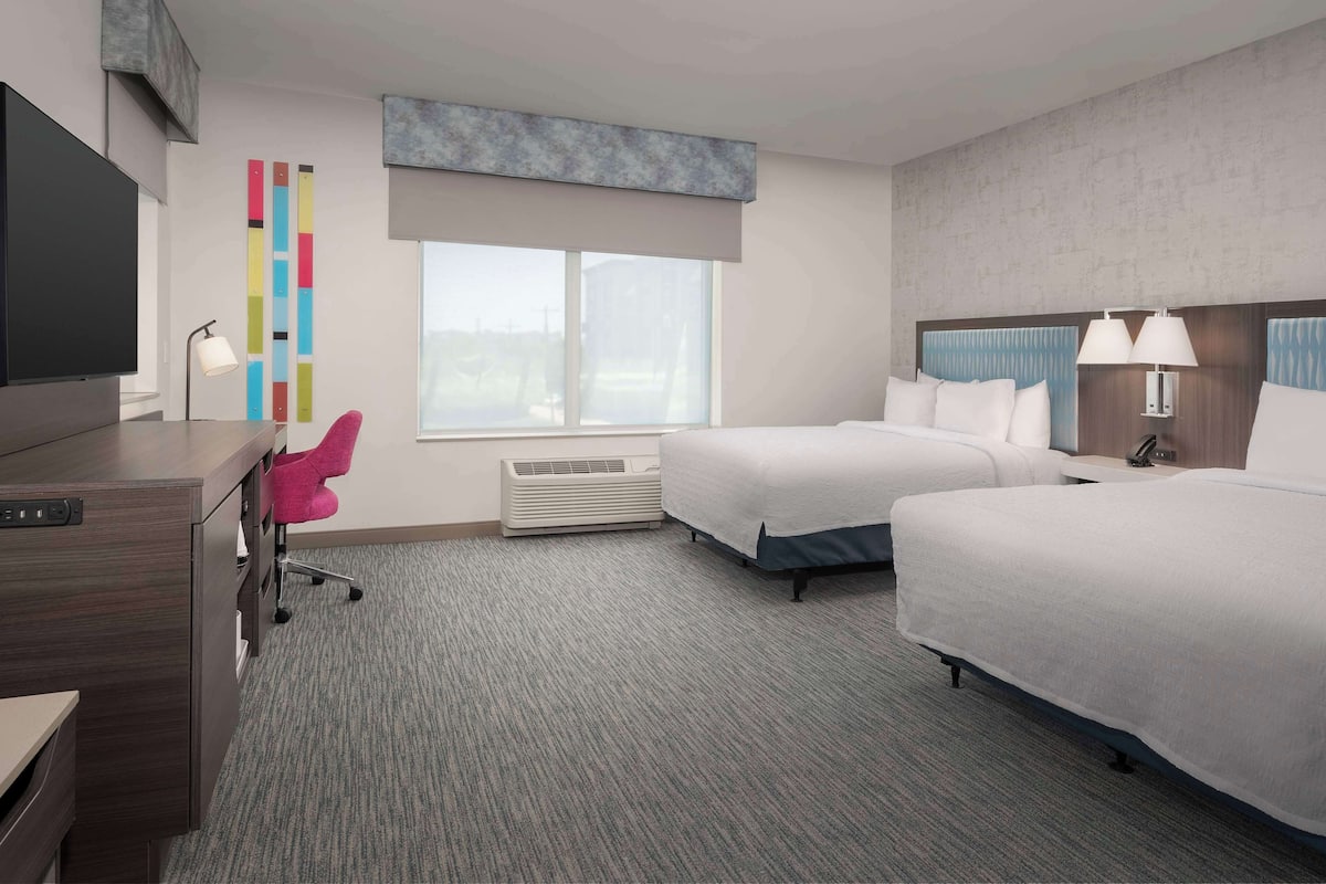 A hotel room with two beds, a desk, a TV, a desk lamp, a chair, a window, and a wall with a colorful vertical design.