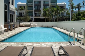 Une piscine entourée d’un mur blanc et de palmiers, avec des chaises longues et un bain de soleil à proximité.