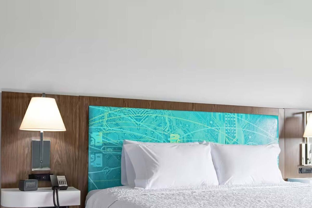 A hotel room with a bed, a nightstand with a lamp, a phone, and a wall with a circuit board design.