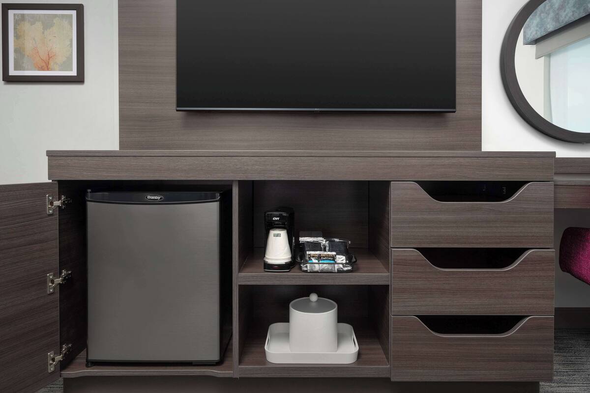A hotel room with a built-in minibar, a flat-screen TV, a coffee maker, and a small seating area with a round mirror.
