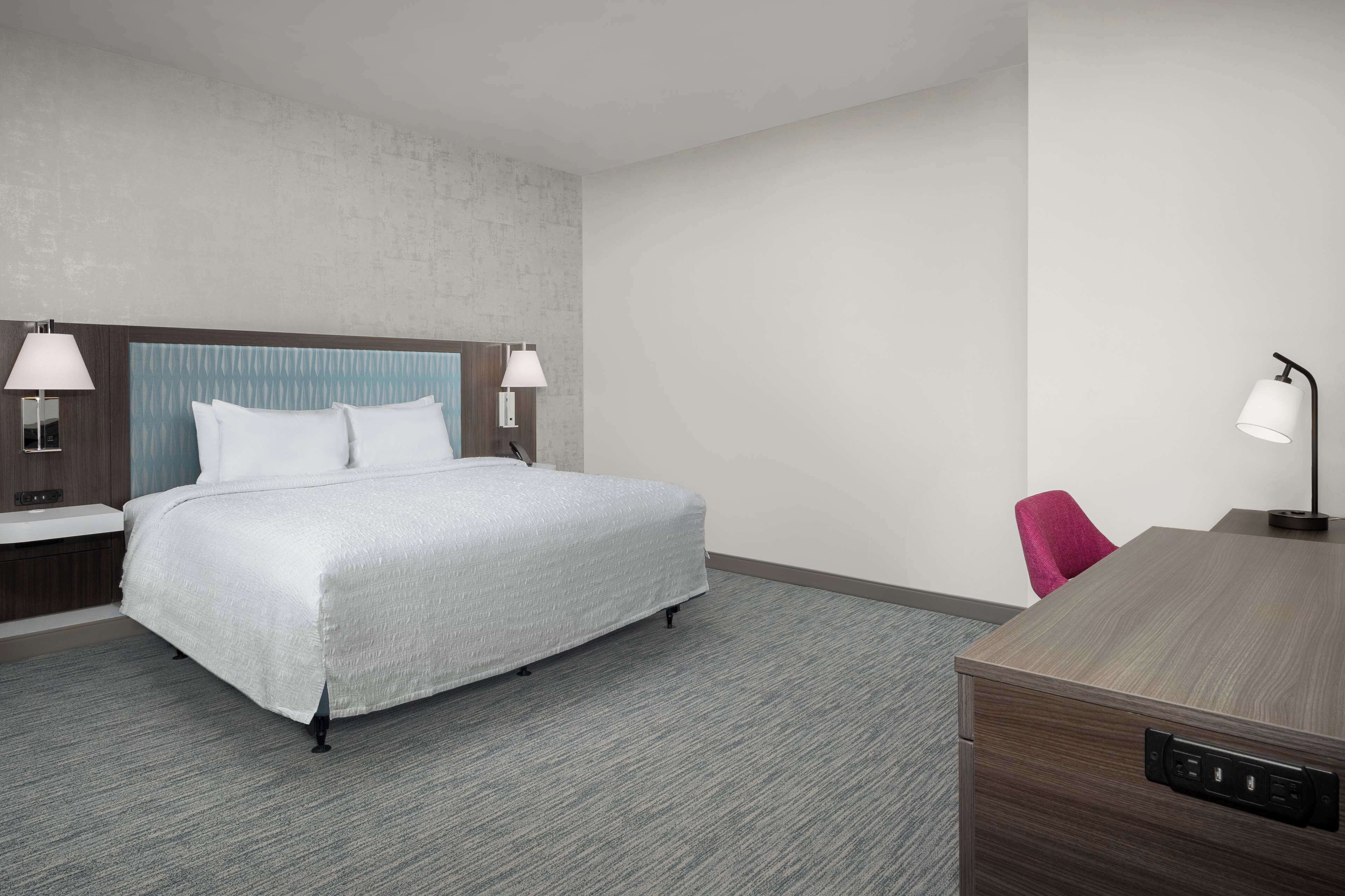 A modern hotel room with a large bed, two bedside lamps, a desk with a lamp, and a chair.