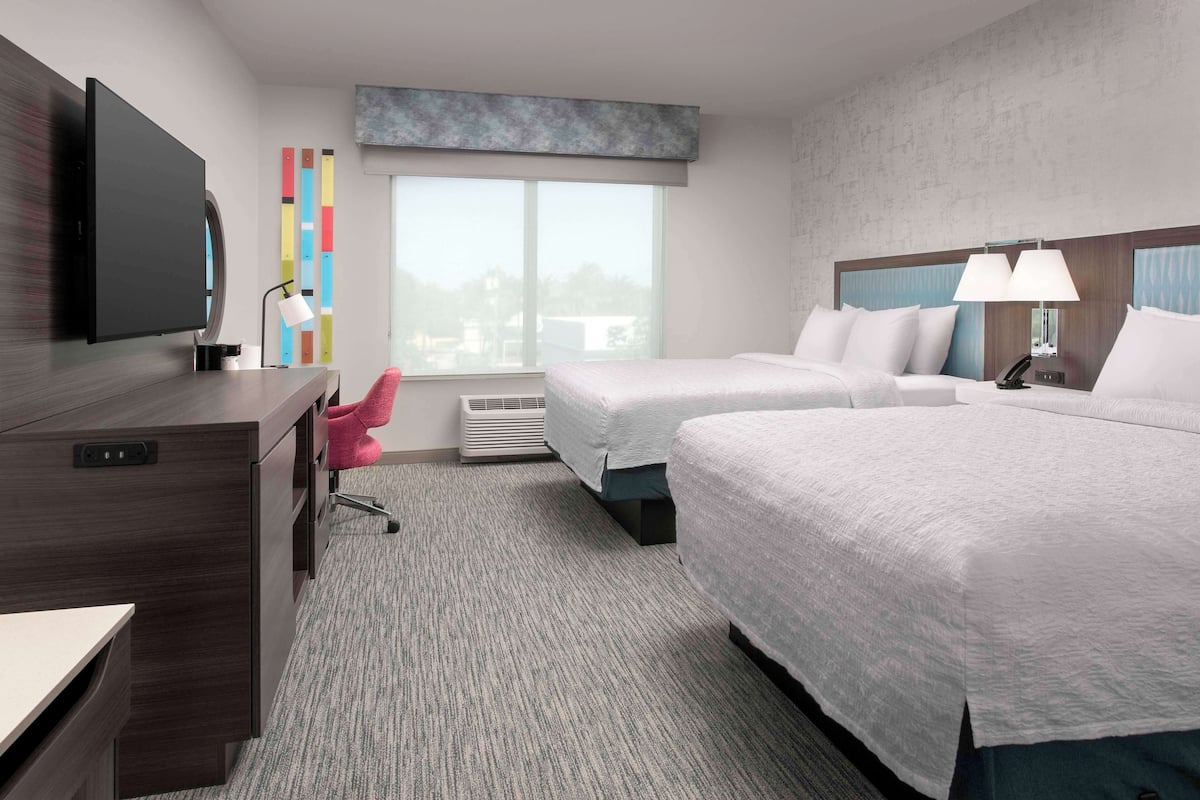 A hotel room with two beds, a desk, a TV, a window, and a colorful wall decoration.