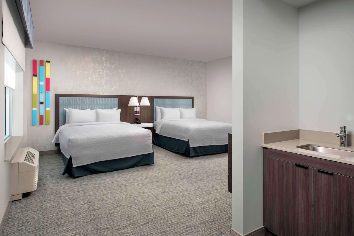 A hotel room with two beds, a small bedside table with lamps, a built-in sink, and a window.
