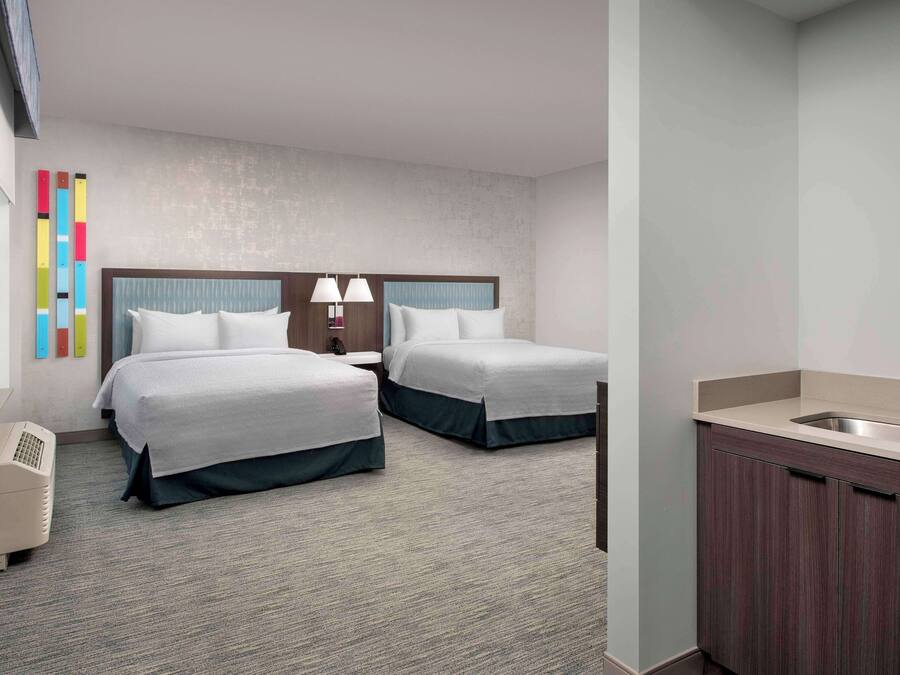 A hotel room with two beds, a small bedside table with lamps, a built-in sink, and a window.
