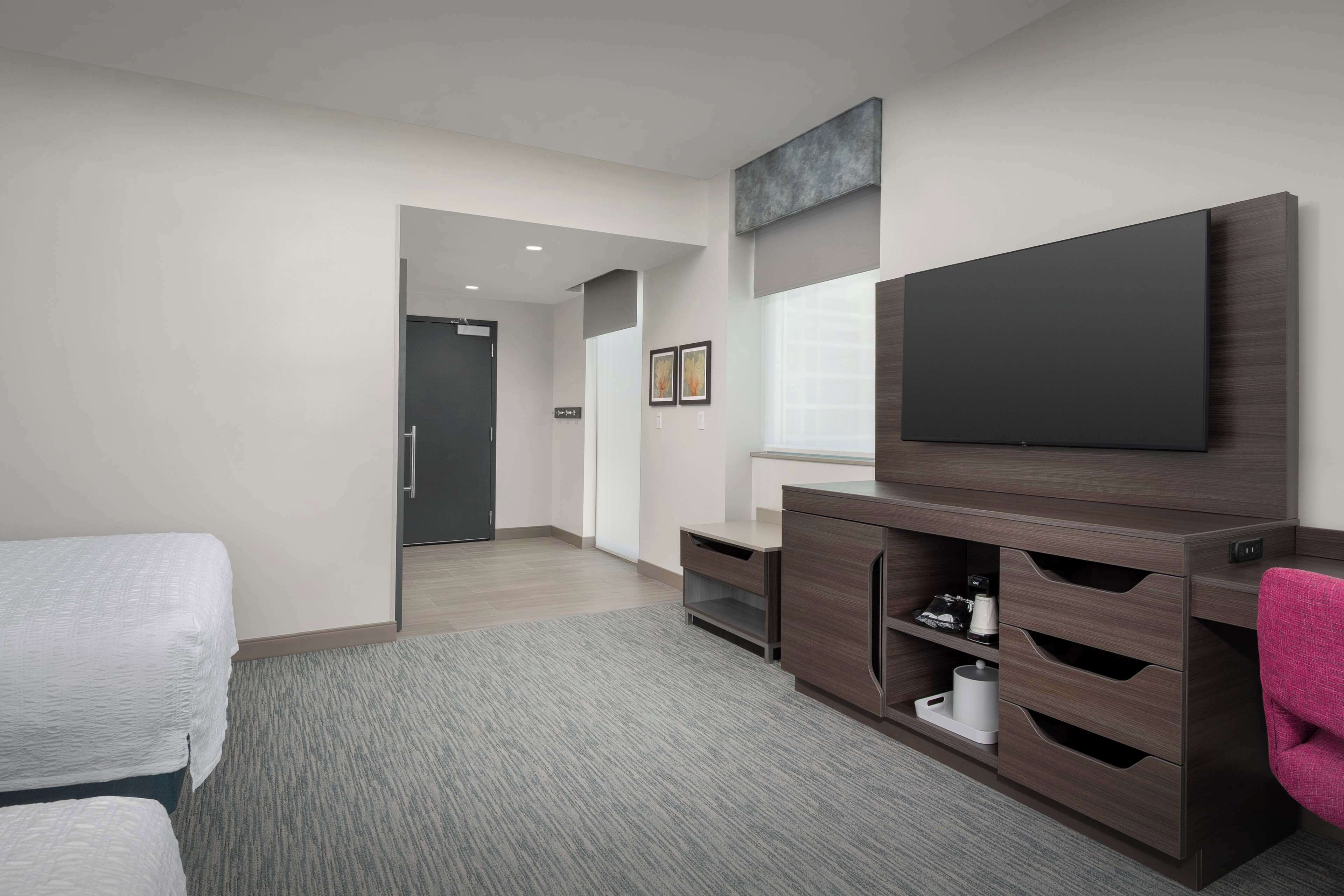 A modern hotel room with a large flat-screen TV, a wooden entertainment unit with storage, a bed with white bedding, and a carpeted floor.