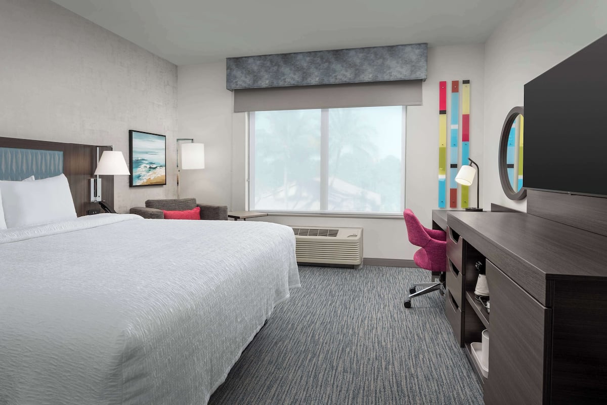 A hotel room with a large bed, a desk, a chair, a window with a view, and a wall with colorful decor.