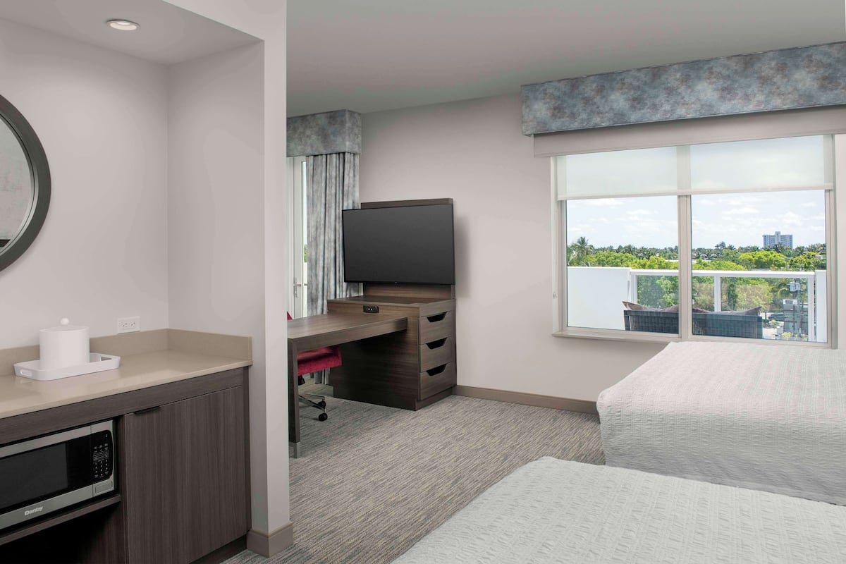 A hotel room with a desk, a flat-screen TV, a bed, and a large window offering a city view.