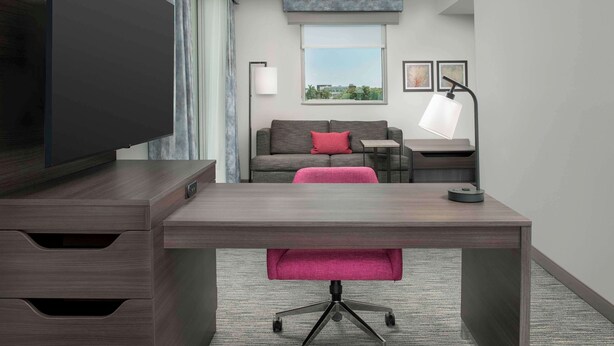A modern hotel room with a large flat-screen TV, a desk with a chair, a sofa with a red pillow, a lamp, and a window with a view of greenery.