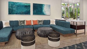 A modern living room with a teal sectional sofa, two large abstract paintings, a dining area with a glass table, and a view of greenery through large windows.