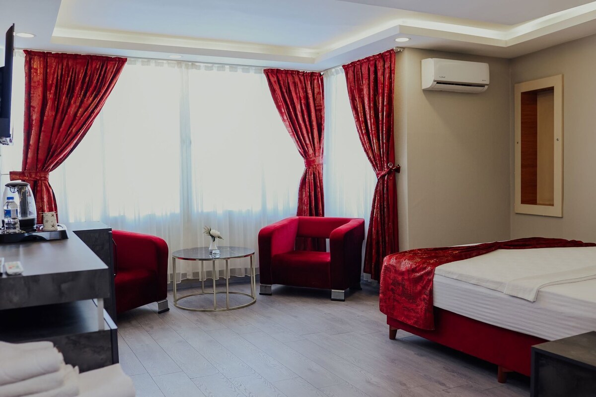 A hotel room with a bed, two red armchairs, a small round table, a coffee maker, and a view of the city through large windows with red curtains.